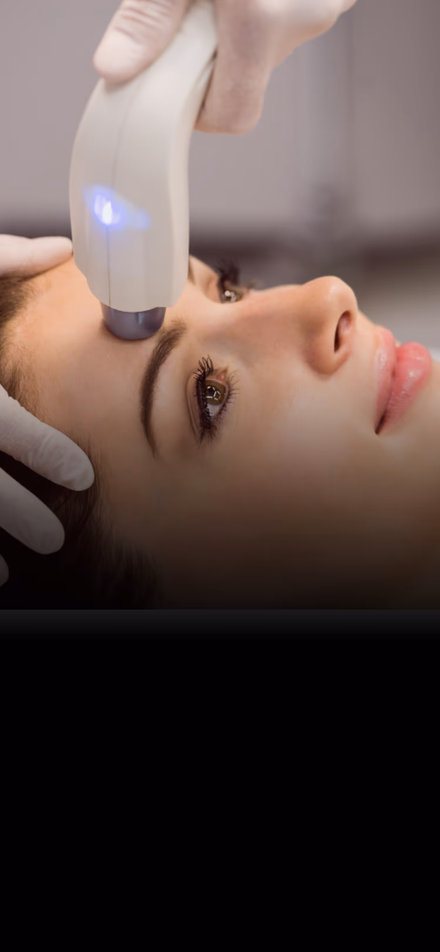 Close-up of a woman receiving a facial treatment with a handheld device emitting blue light on her forehead.