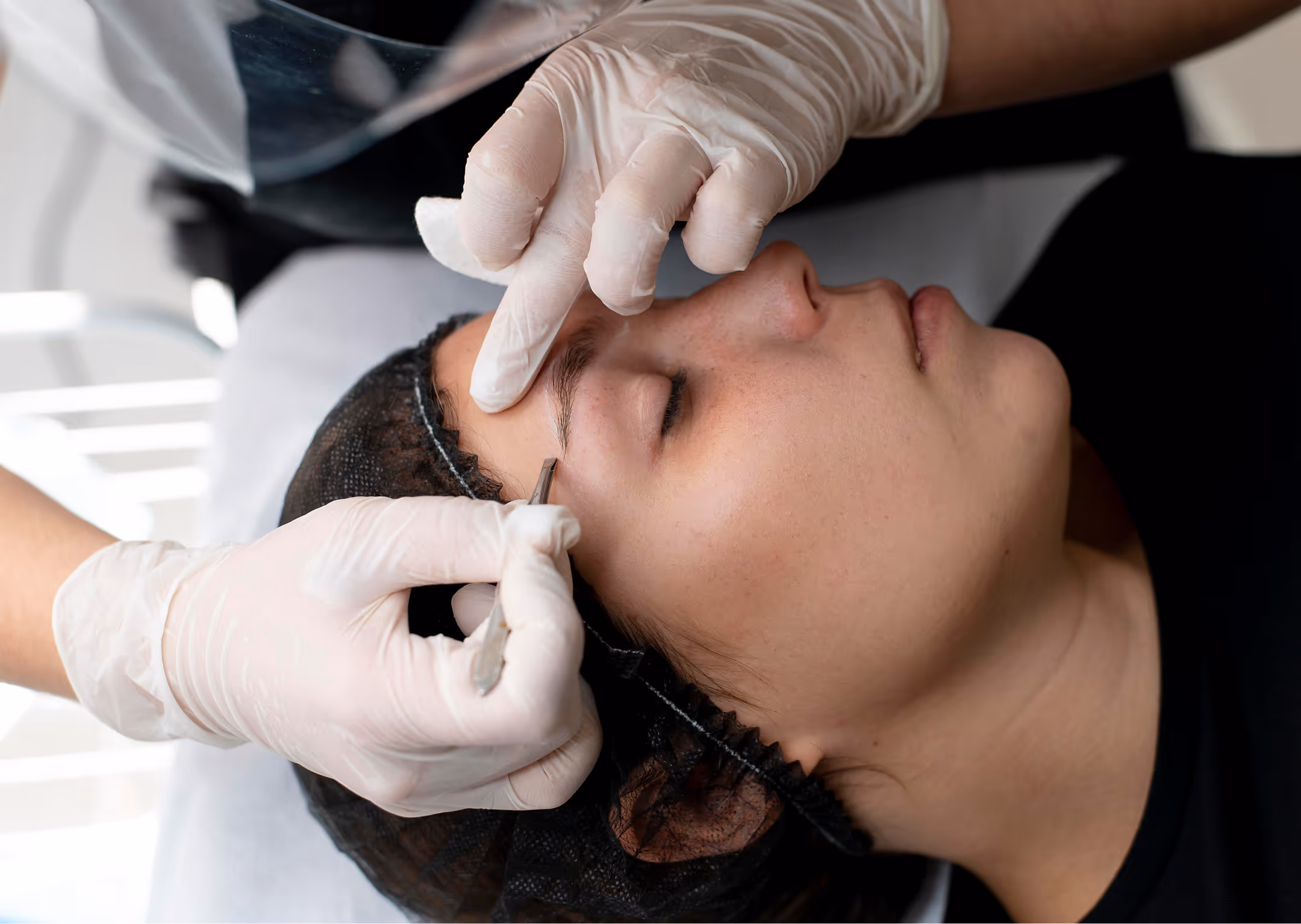 Gloved hands performing microblading on a woman’s eyebrow as she lies with eyes closed.