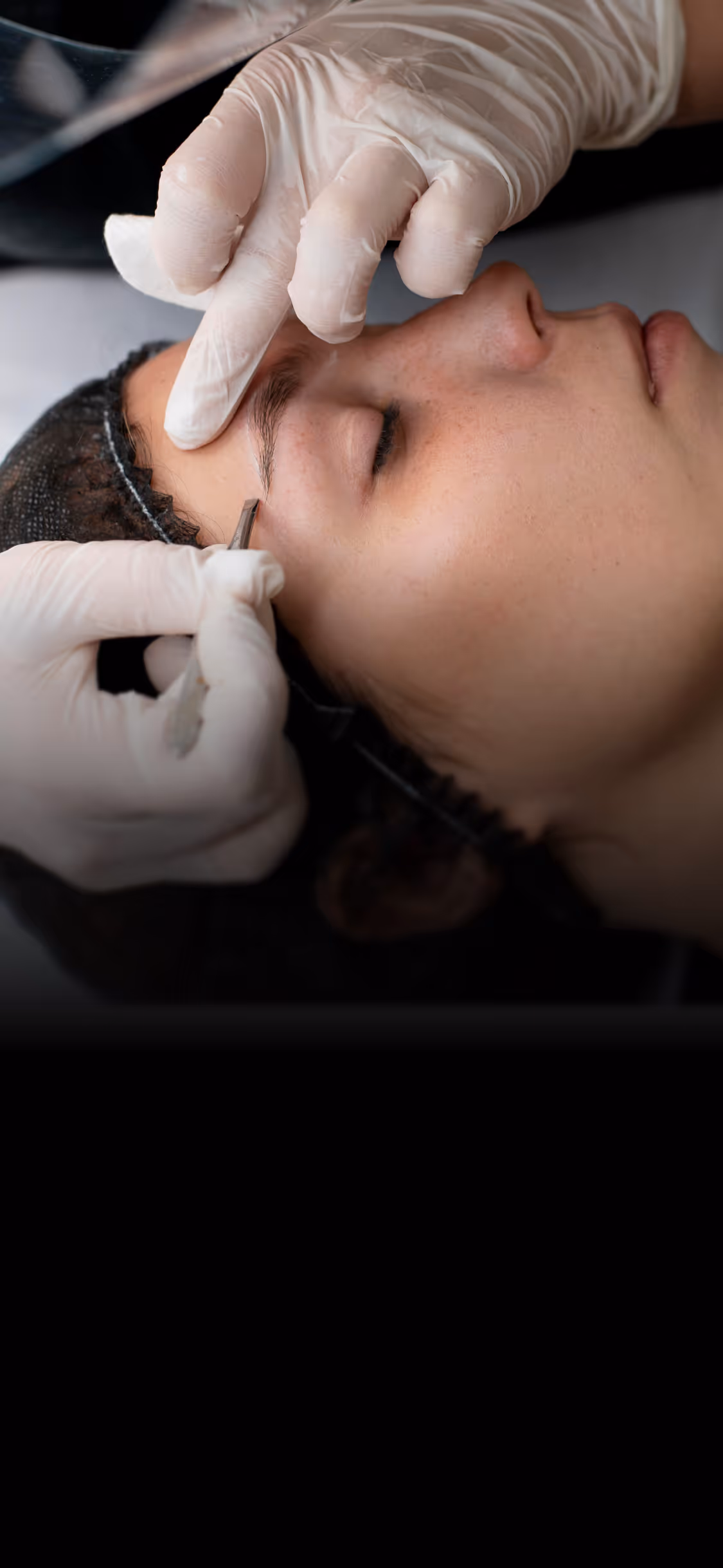 Hands in gloves shaping a woman's eyebrow with tweezers during a beauty treatment.