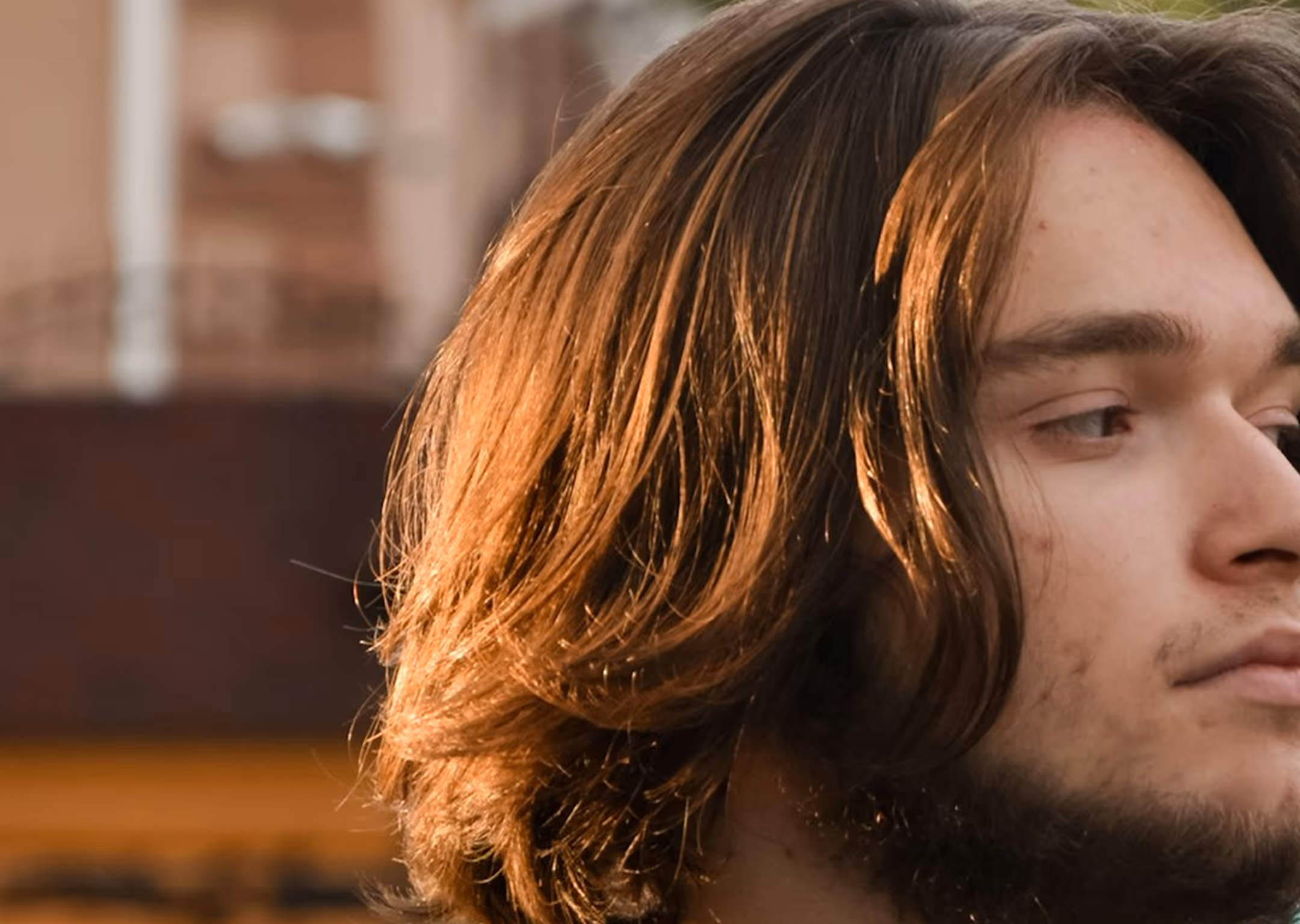 Close-up of a young man with medium long brown hair and beard looking to the side outdoors.