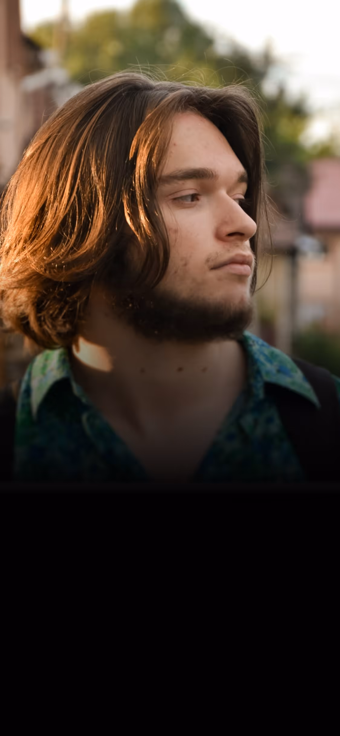 Young man with medium-length brown hair and beard looking to the side outdoors in natural light.