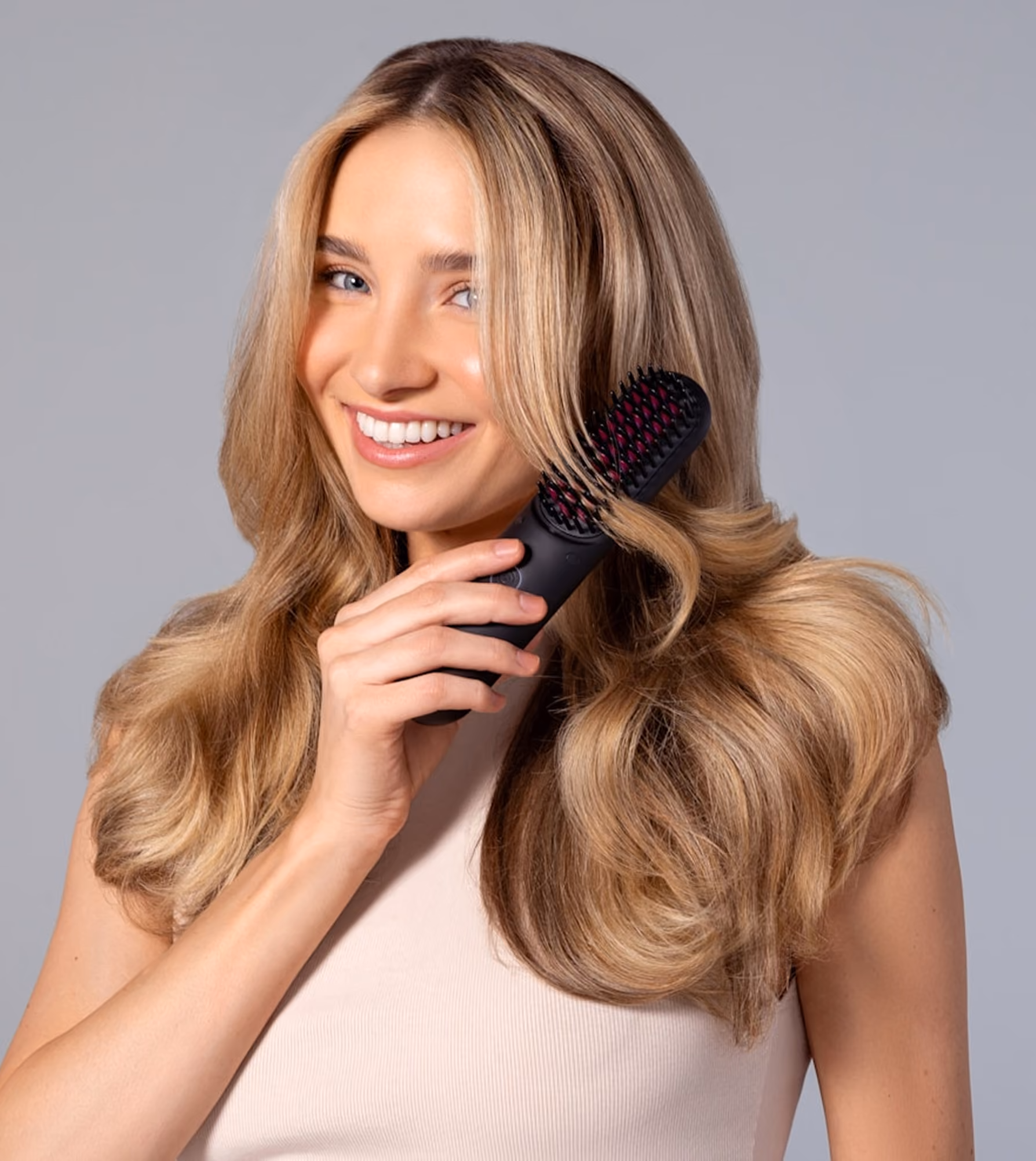 Smiling woman with long blonde hair using a black heated hair straightening brush.