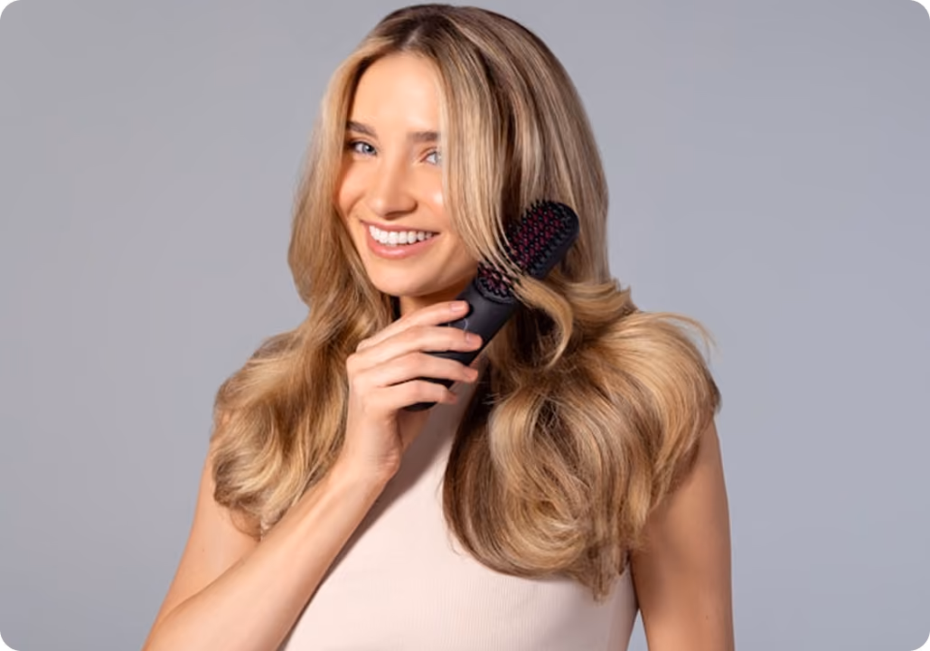 Smiling woman with long blonde hair using a black hairbrush against a gray background.