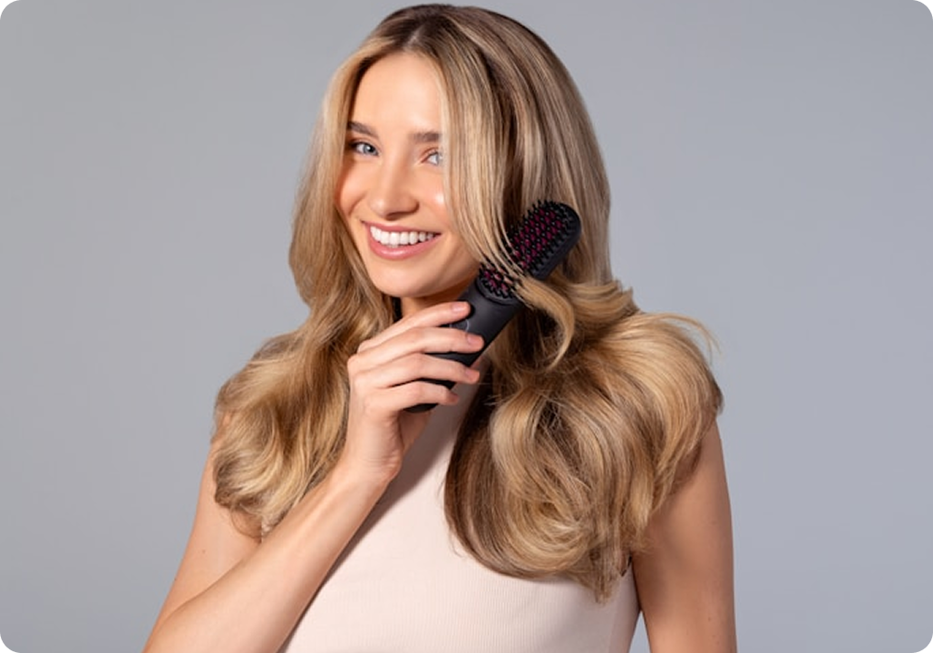 Smiling woman with long blonde hair using a black hairbrush against a gray background.