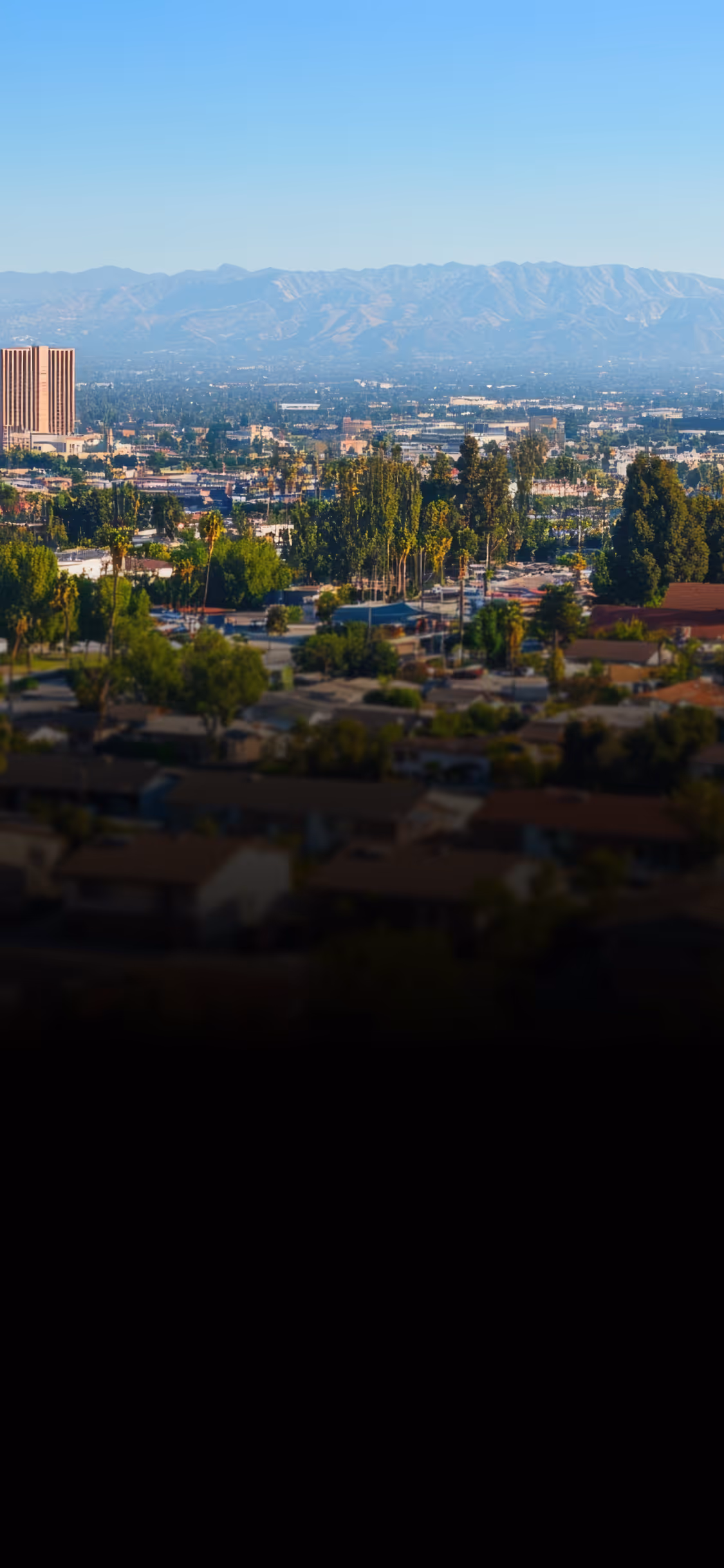 Cityscape with tall buildings, trees, houses, and mountains under a clear blue sky.