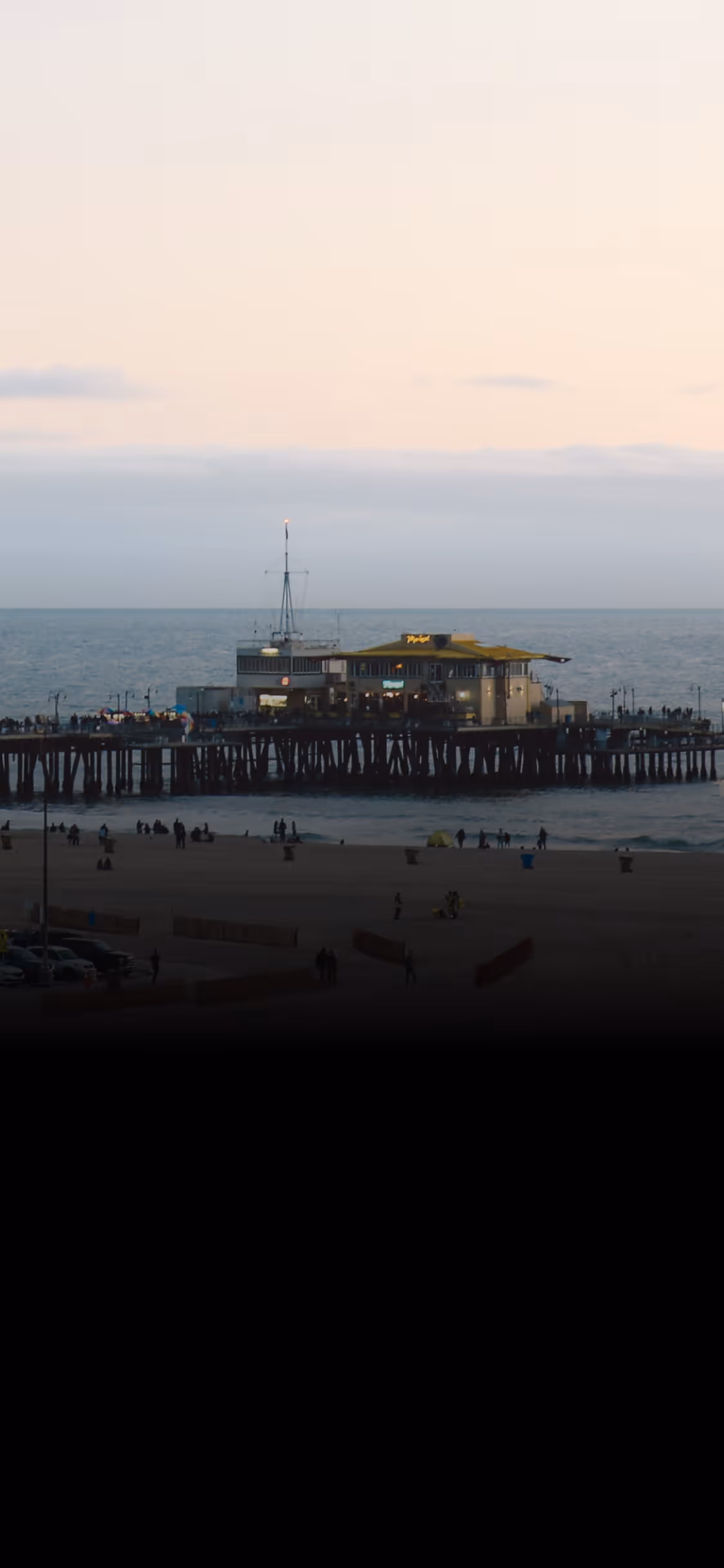 Crowded pier with building and people on beach at sunset under cloudy sky by the ocean.