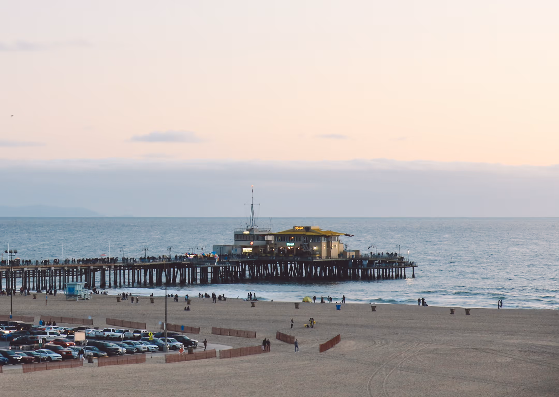 Pier with a building extends into the ocean at sunset, with beach, people, and parked cars nearby.