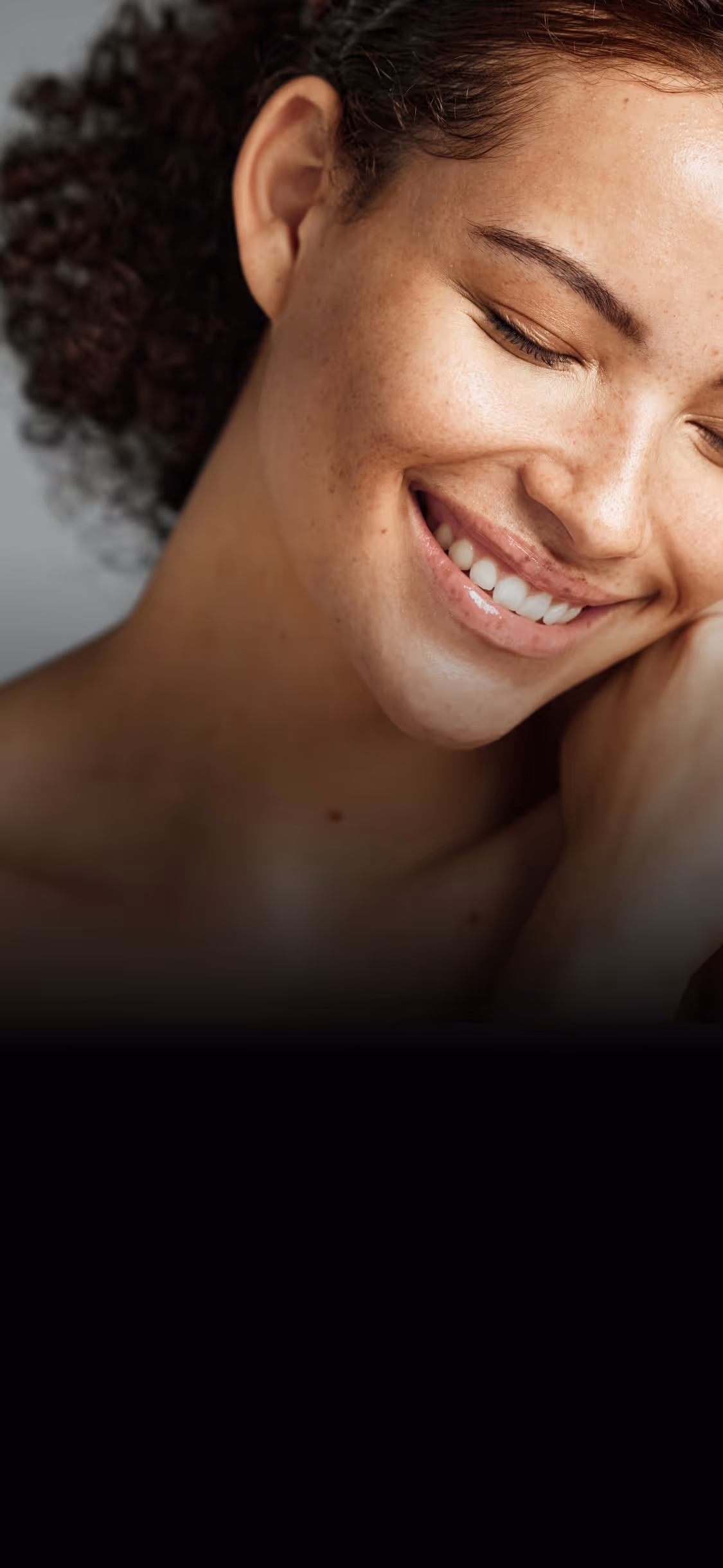 Close-up of a smiling woman with curly hair and closed eyes resting her face on her hand.