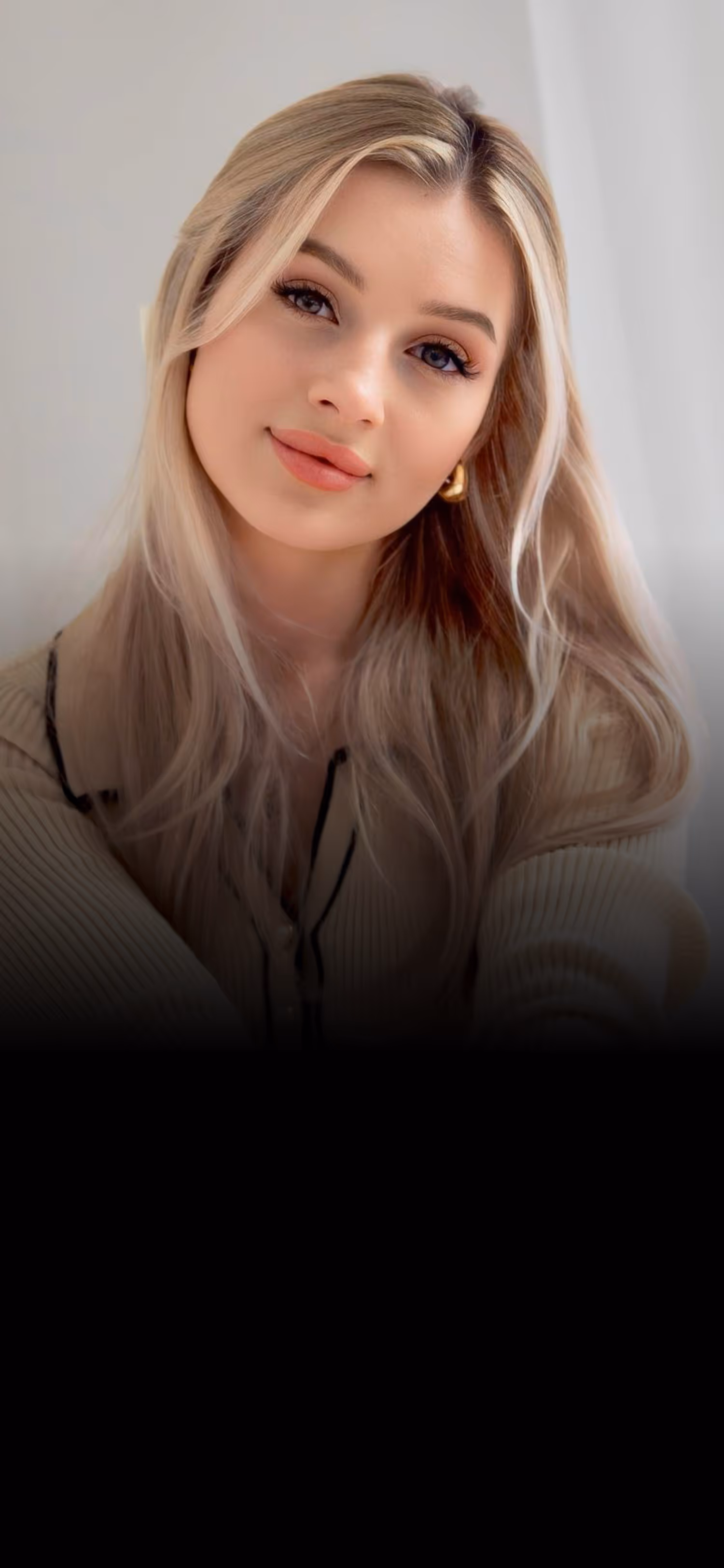 Young woman with long blonde hair wearing gold hoop earrings and a beige sweater smiles softly.