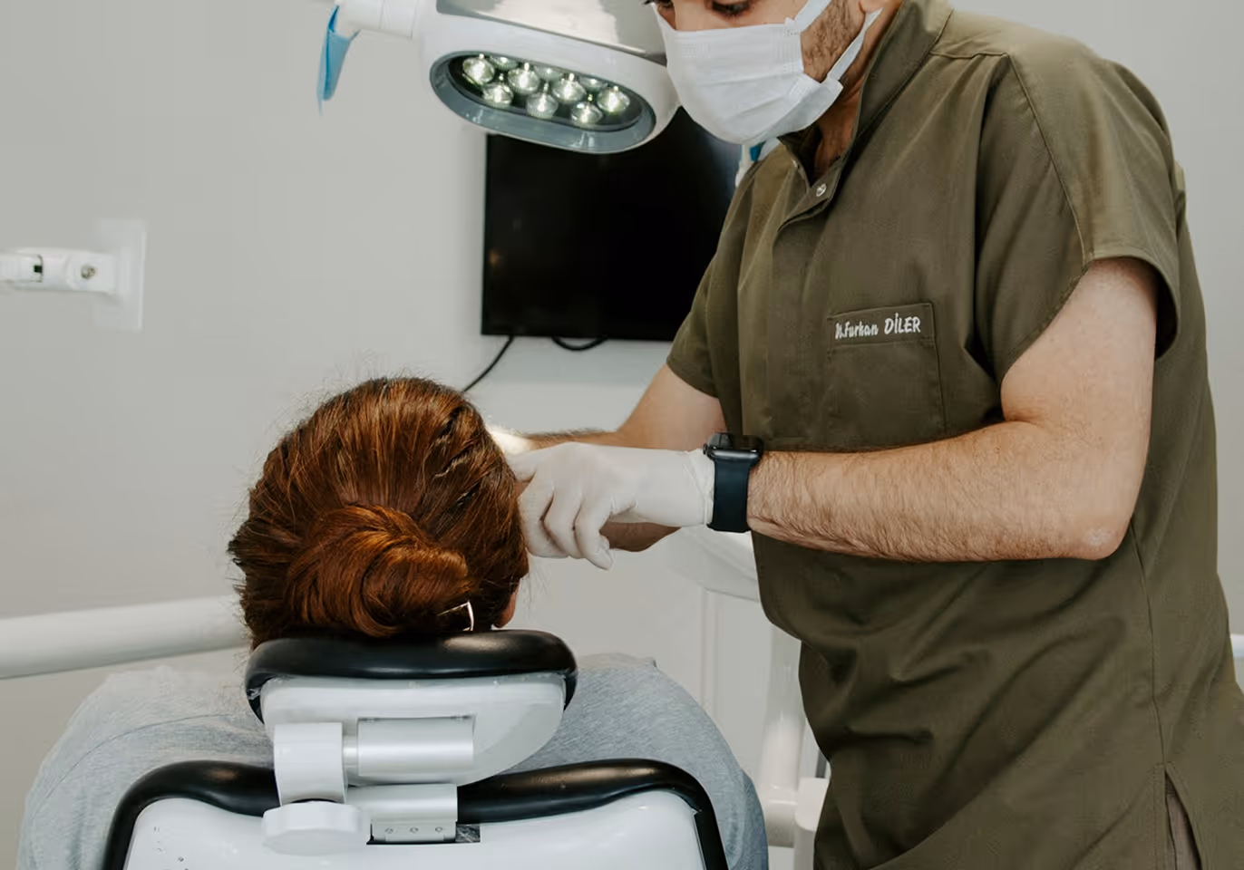 Dentist wearing mask and gloves examining a patient seated in a dental chair under a light.