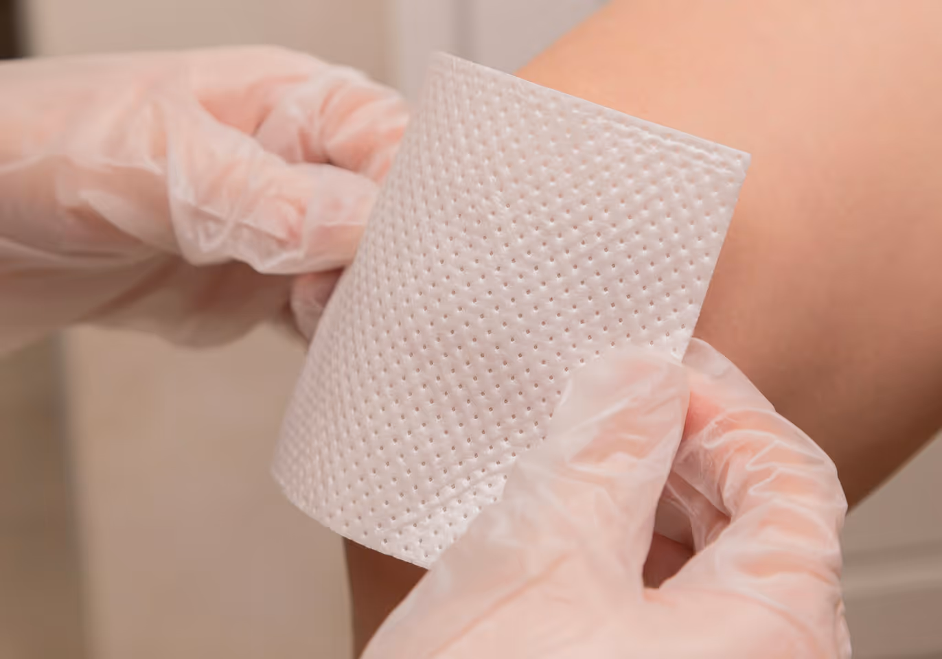 Hands in medical gloves applying a textured adhesive bandage to a bare knee.