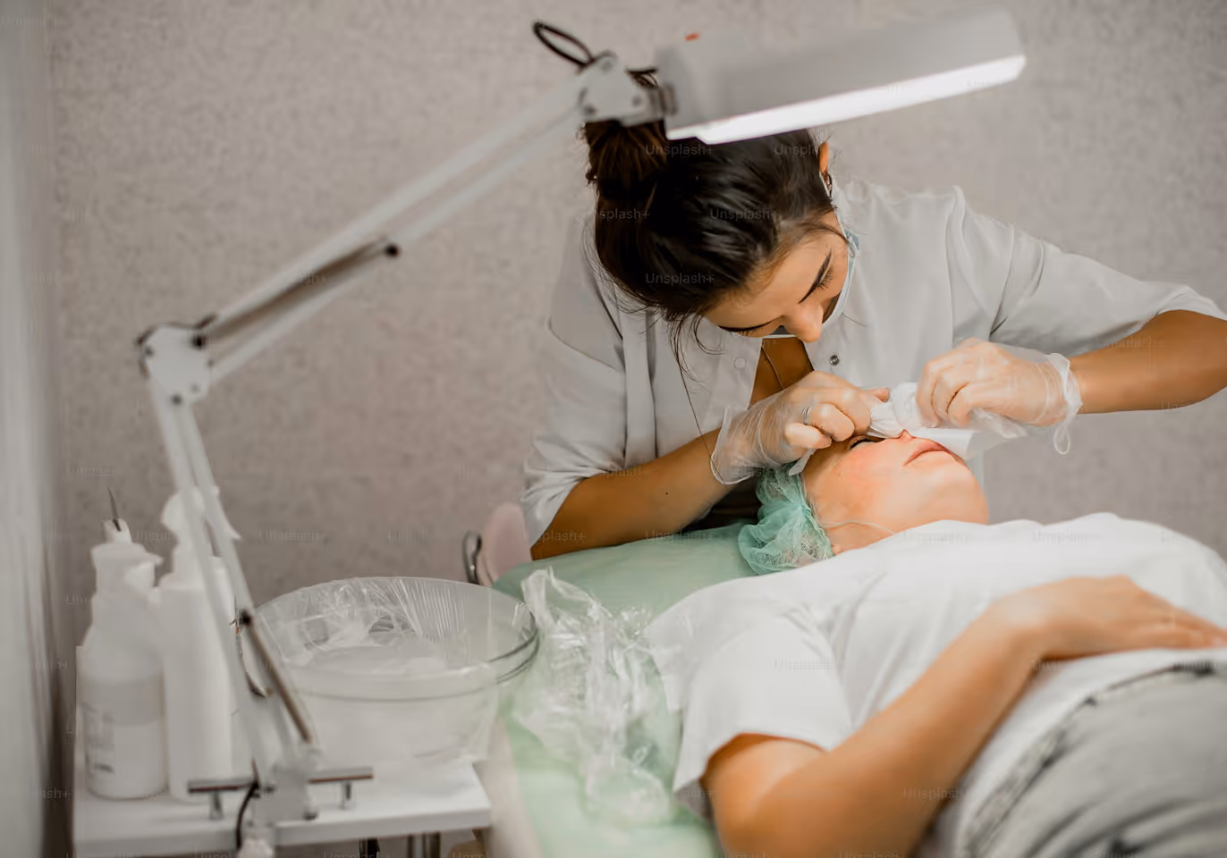 A beautician wearing gloves cleans a client's face under a bright lamp in a spa treatment room.