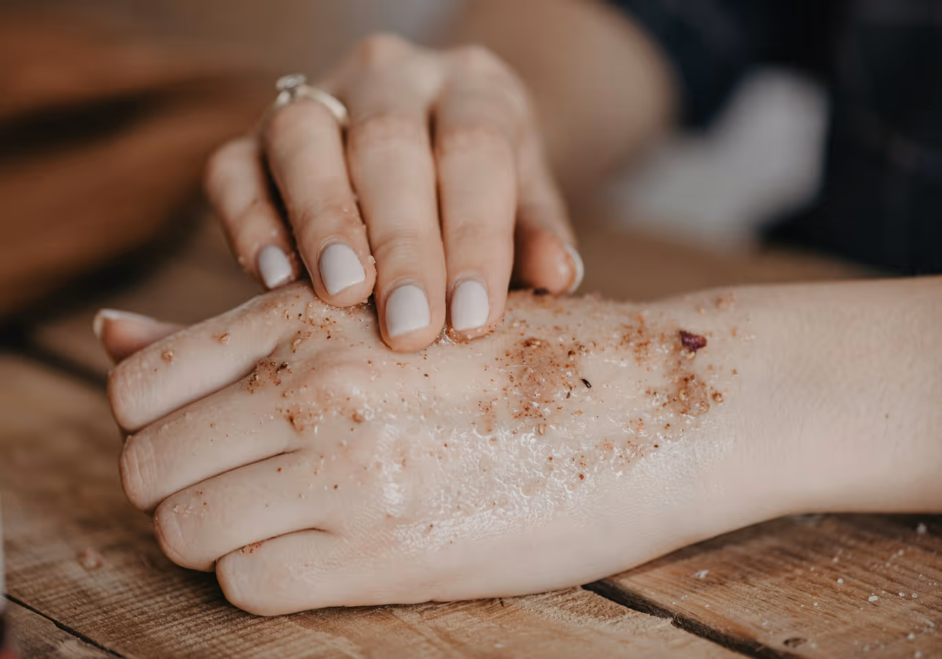 Person rubbing exfoliating scrub with granules on the back of their hand over wooden surface.
