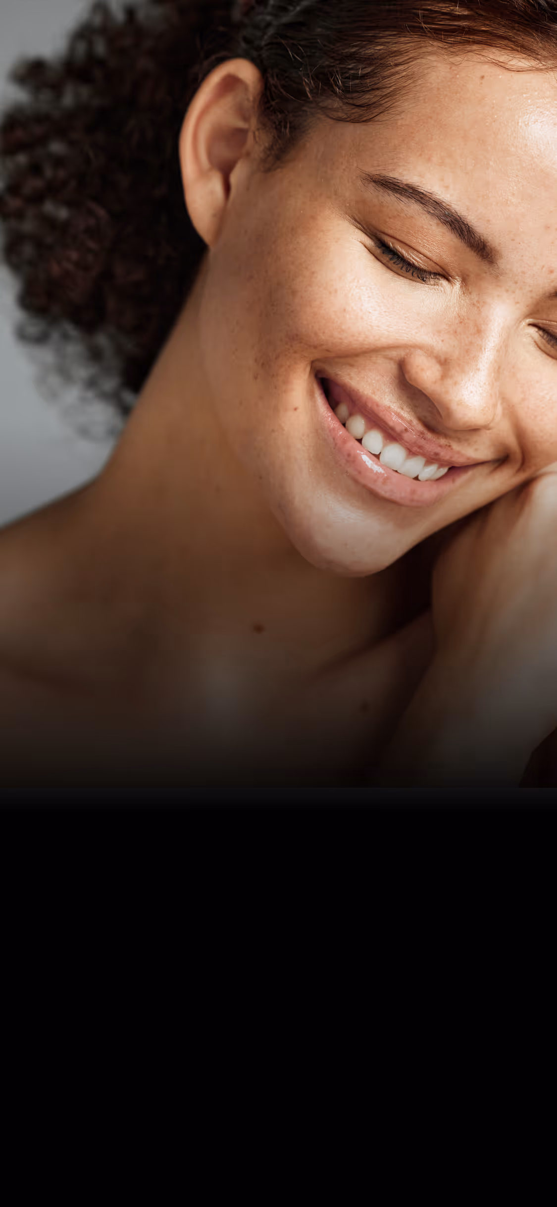 Close-up of a smiling woman with curly hair and freckles, eyes gently closed.