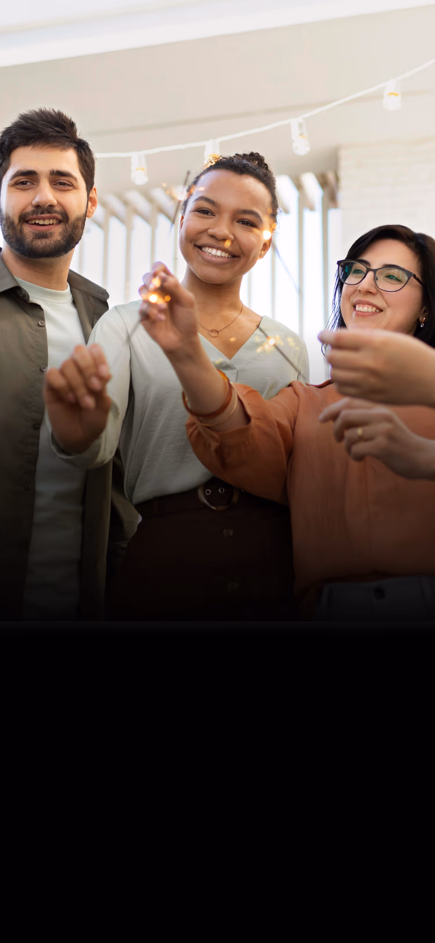 Three smiling people holding lit sparklers indoors with string lights in the background.