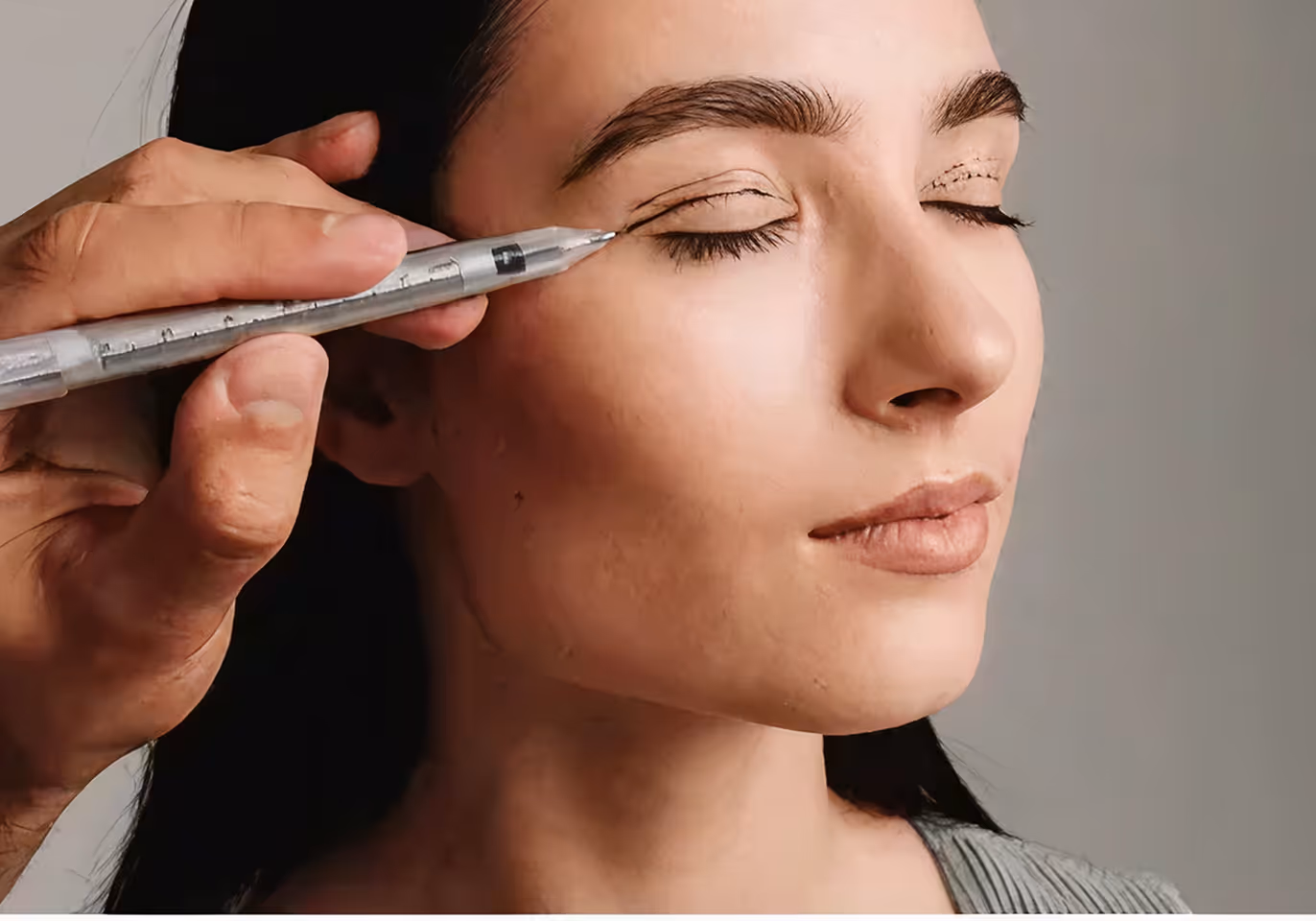 Close-up of a woman with eyes closed as eyeliner is being drawn on her upper eyelid with a pen.