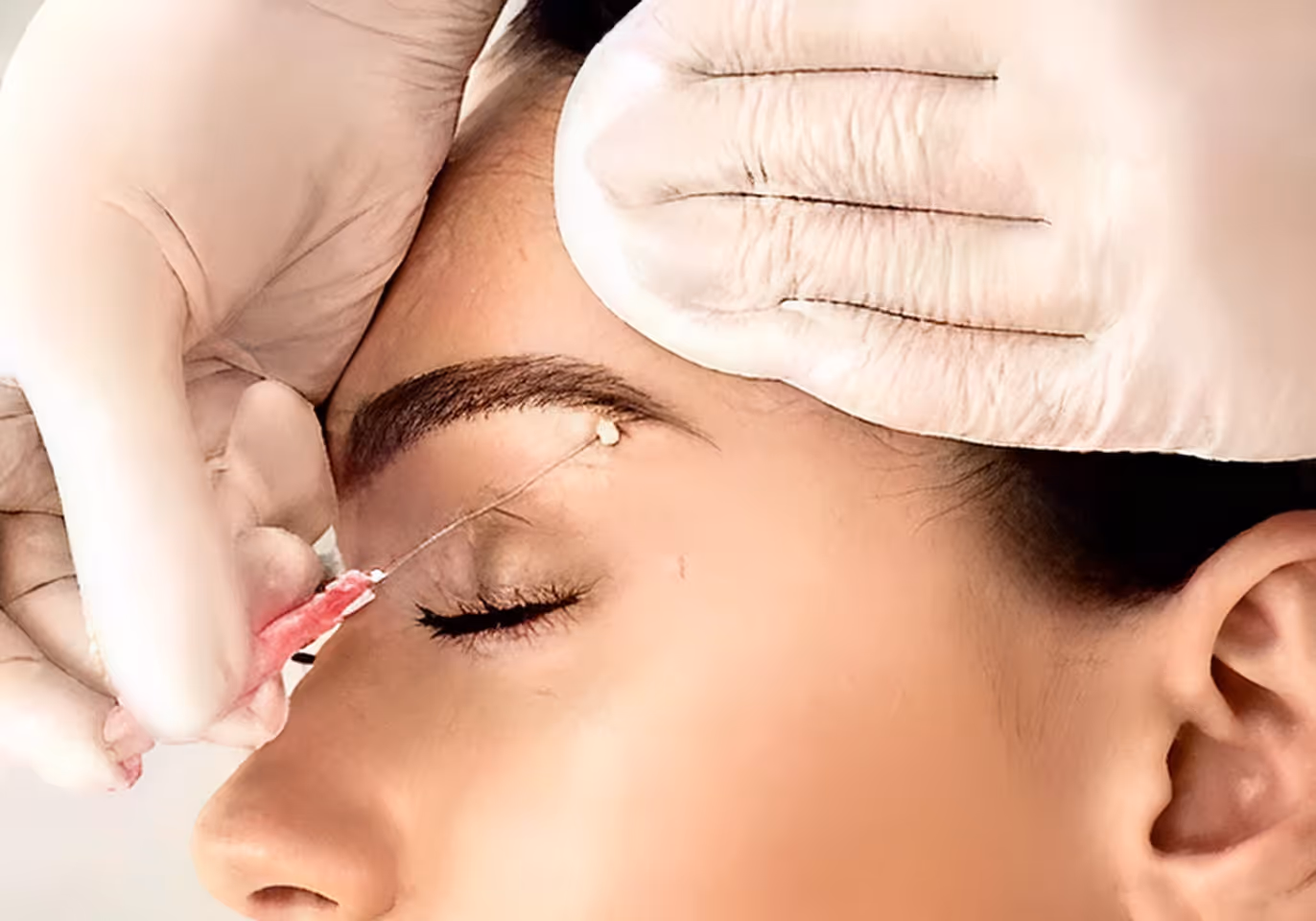 Gloved hands performing an eyebrow threading procedure on a closed eye.