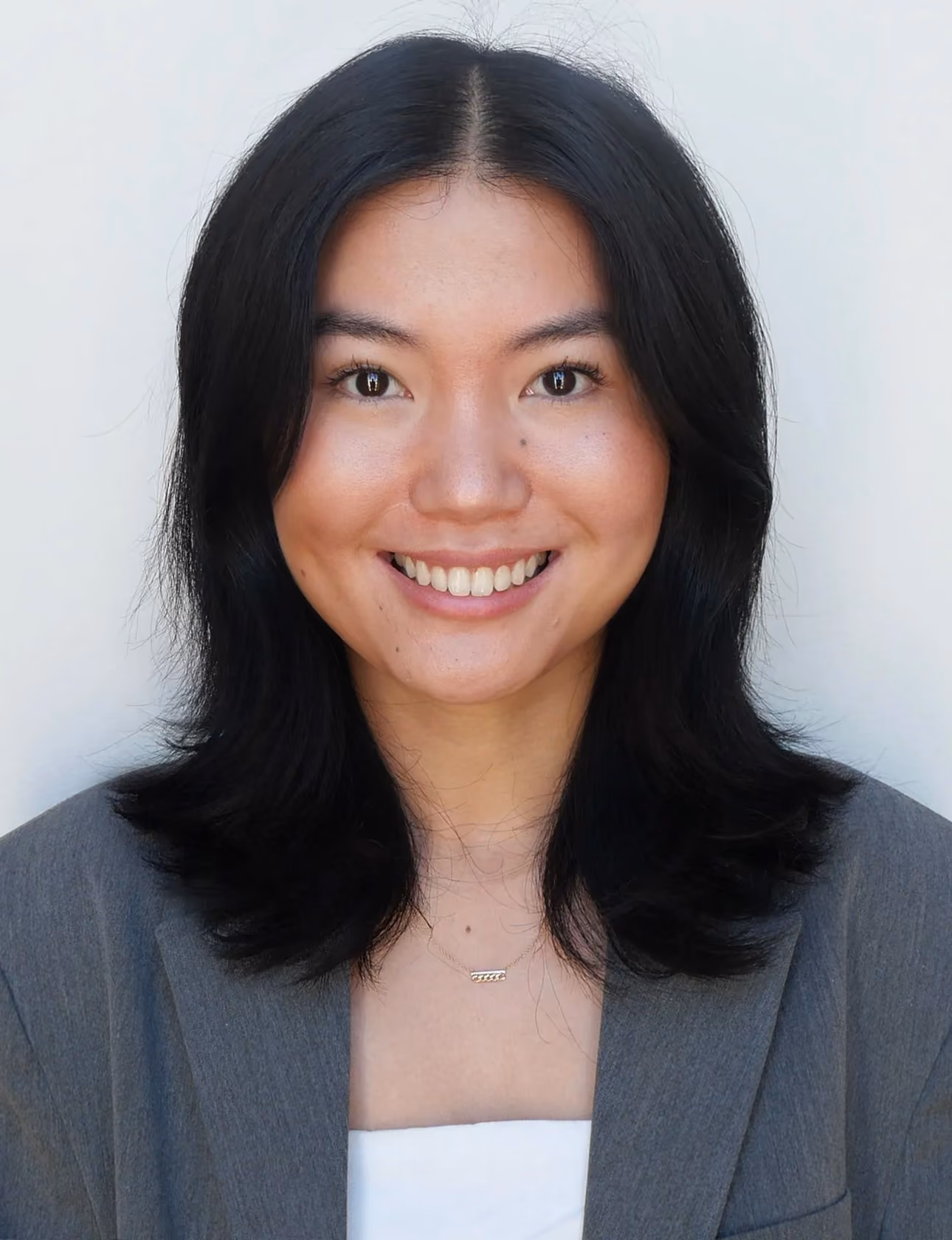 Smiling woman with shoulder-length black hair wearing a gray blazer and white top on a light background.