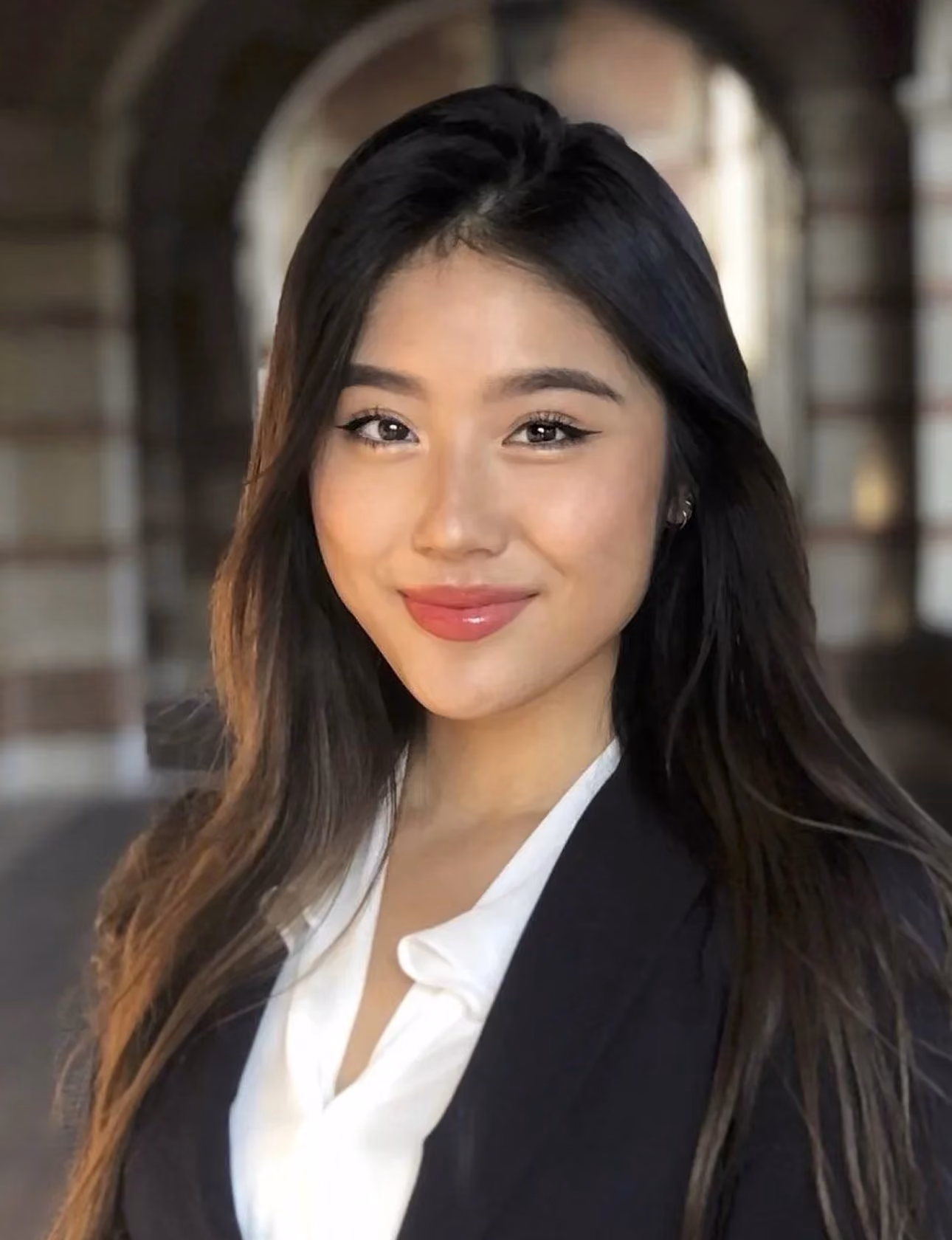 Smiling woman with long dark hair wearing white blouse and black blazer in an arched hallway.