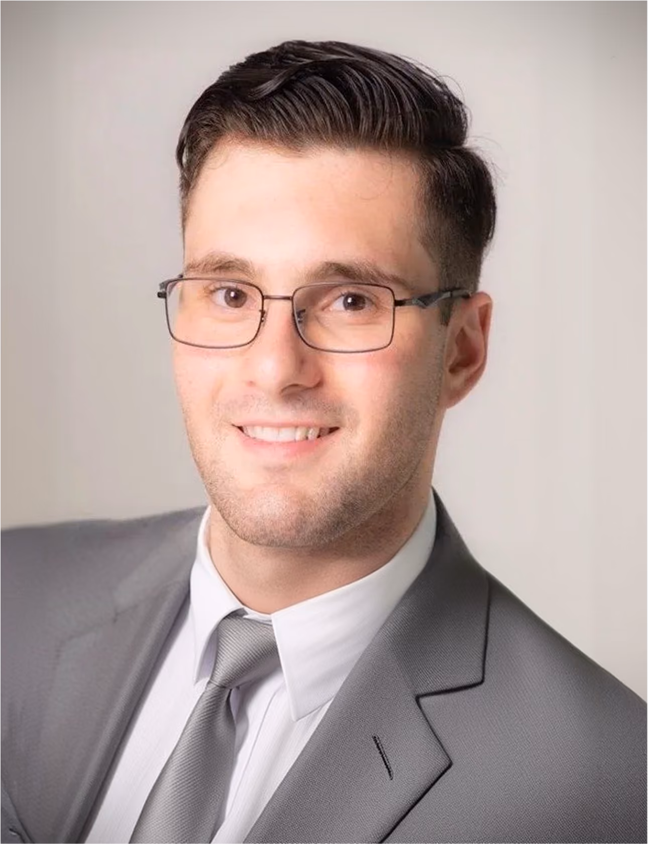 Smiling man with glasses wearing a gray suit, white shirt, and gray tie on a light background.