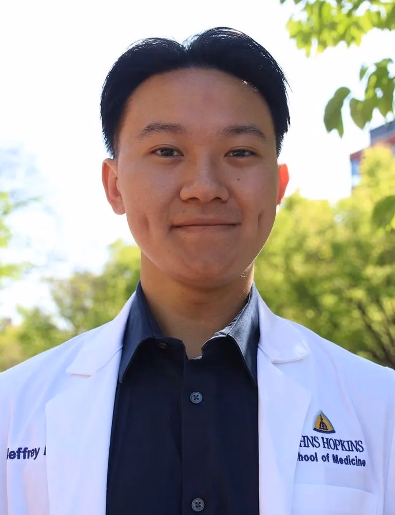 Young man with dimples wearing a white Johns Hopkins School of Medicine coat outdoors.