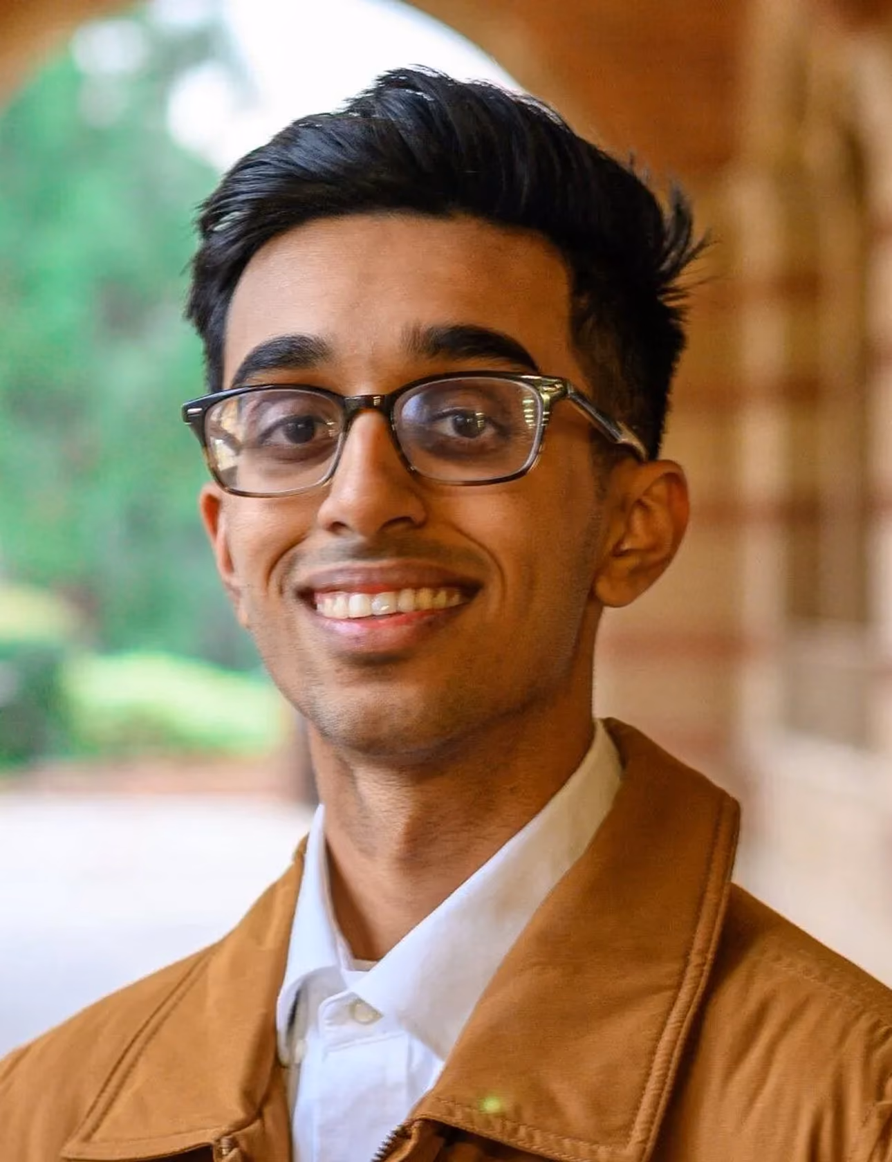 Smiling young man wearing glasses and a brown jacket with a blurred outdoor background.