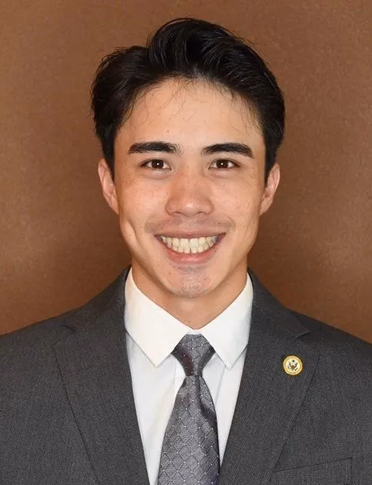 Smiling man wearing a gray suit, white shirt, and patterned gray tie against a brown background.