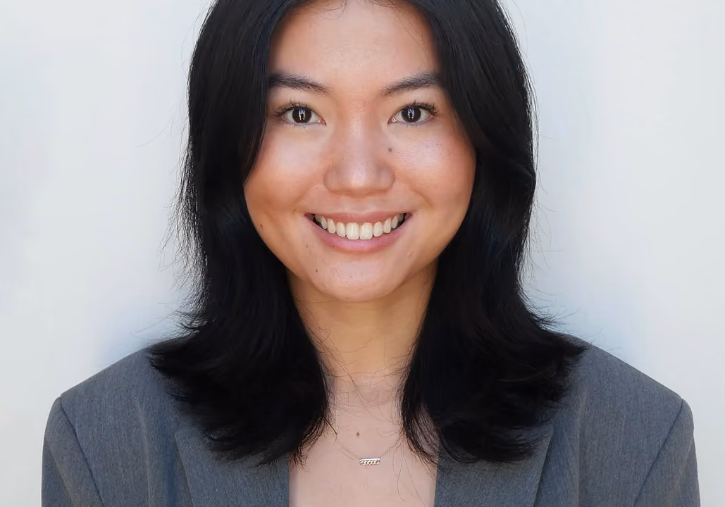 Smiling young woman with black hair wearing a gray blazer against a white background.