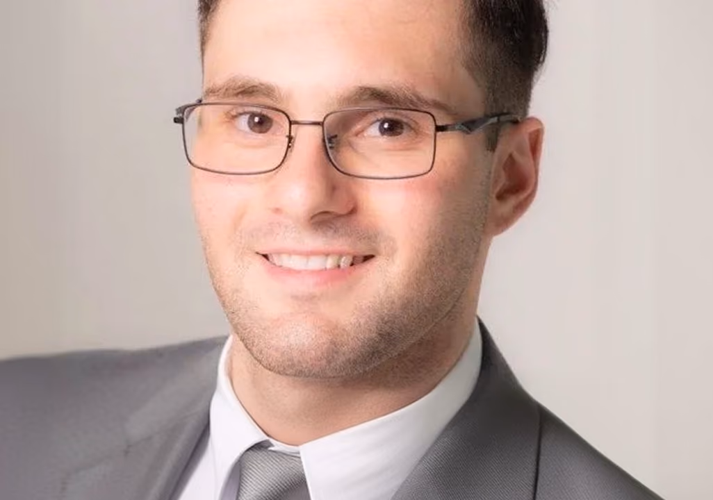 Smiling man wearing glasses, a gray suit, white shirt, and gray tie against a plain background.