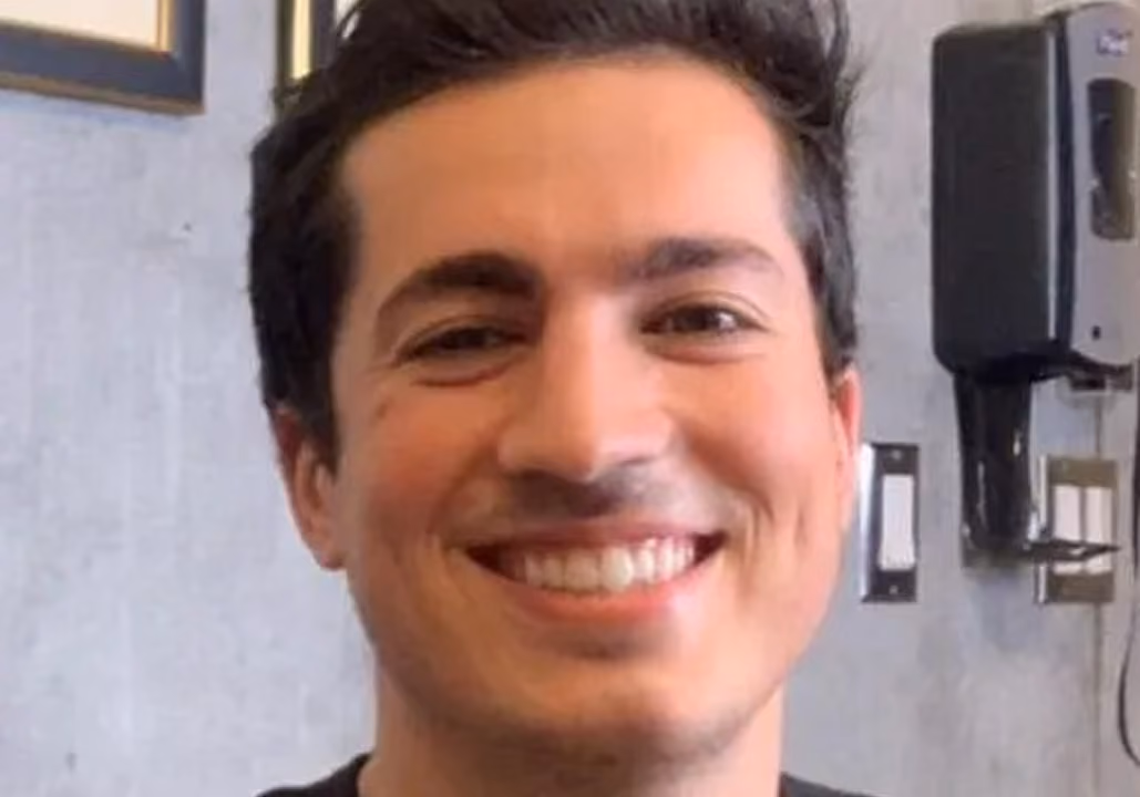 Close-up of a young man smiling against a light gray wall background.