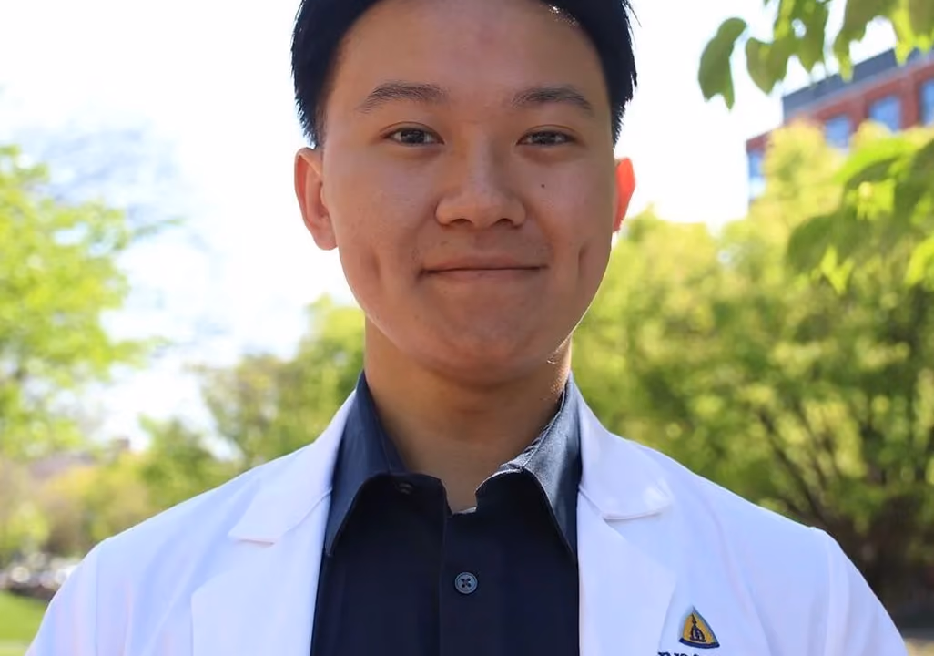 Smiling young man with dimples wearing a white coat and dark shirt outdoors with greenery background.