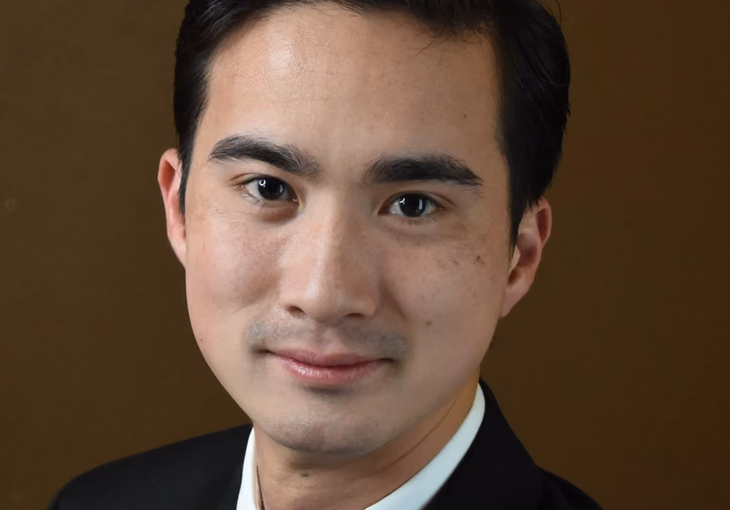 Close-up of a young man with dark hair and brown eyes wearing a black jacket and white shirt.