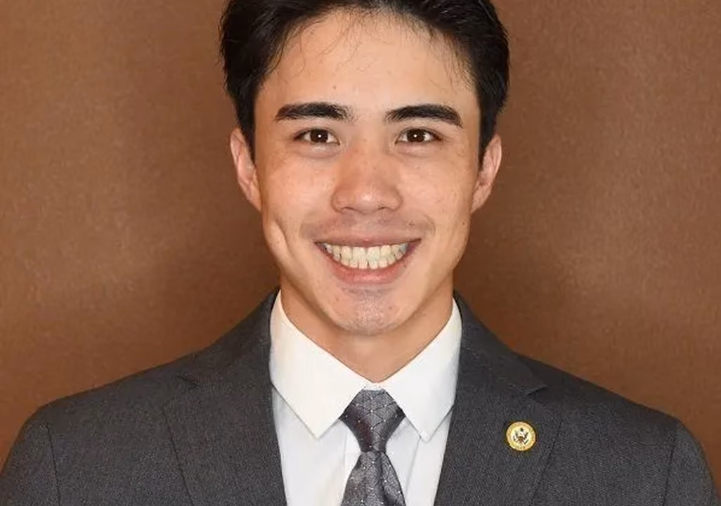 Smiling young man with dark hair wearing a gray suit, white shirt, and patterned tie against brown background.