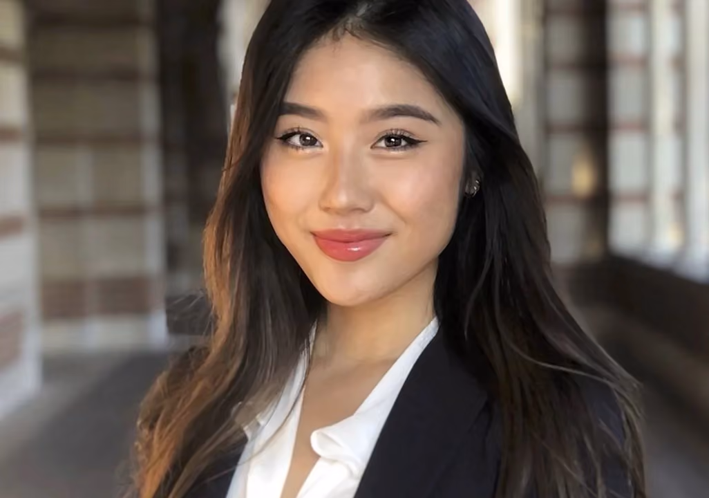 Young woman with long dark hair smiling, wearing a white blouse and black blazer in blurred setting.