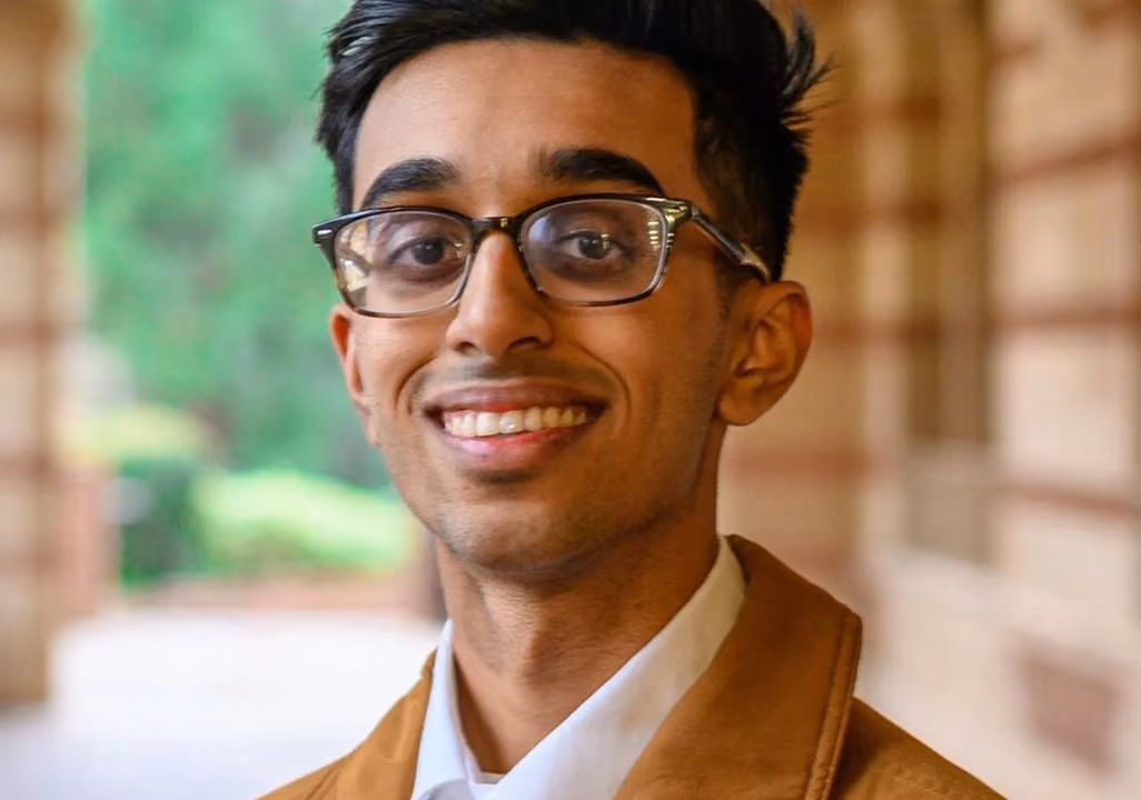 Young man with glasses smiling, wearing a white shirt and brown jacket, blurred background.