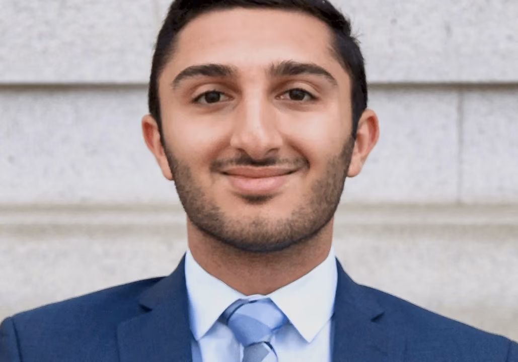 Smiling man with beard wearing a blue suit, white shirt, and blue tie against a stone wall.