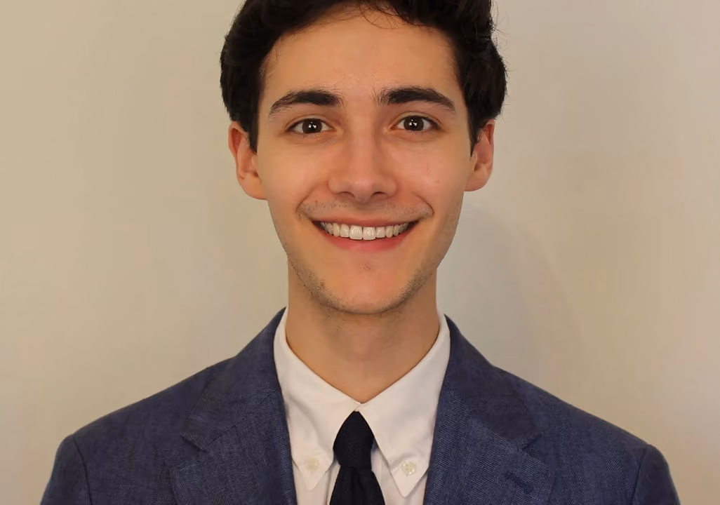 Smiling young man in a blue suit jacket, white shirt, and black tie against a plain background.