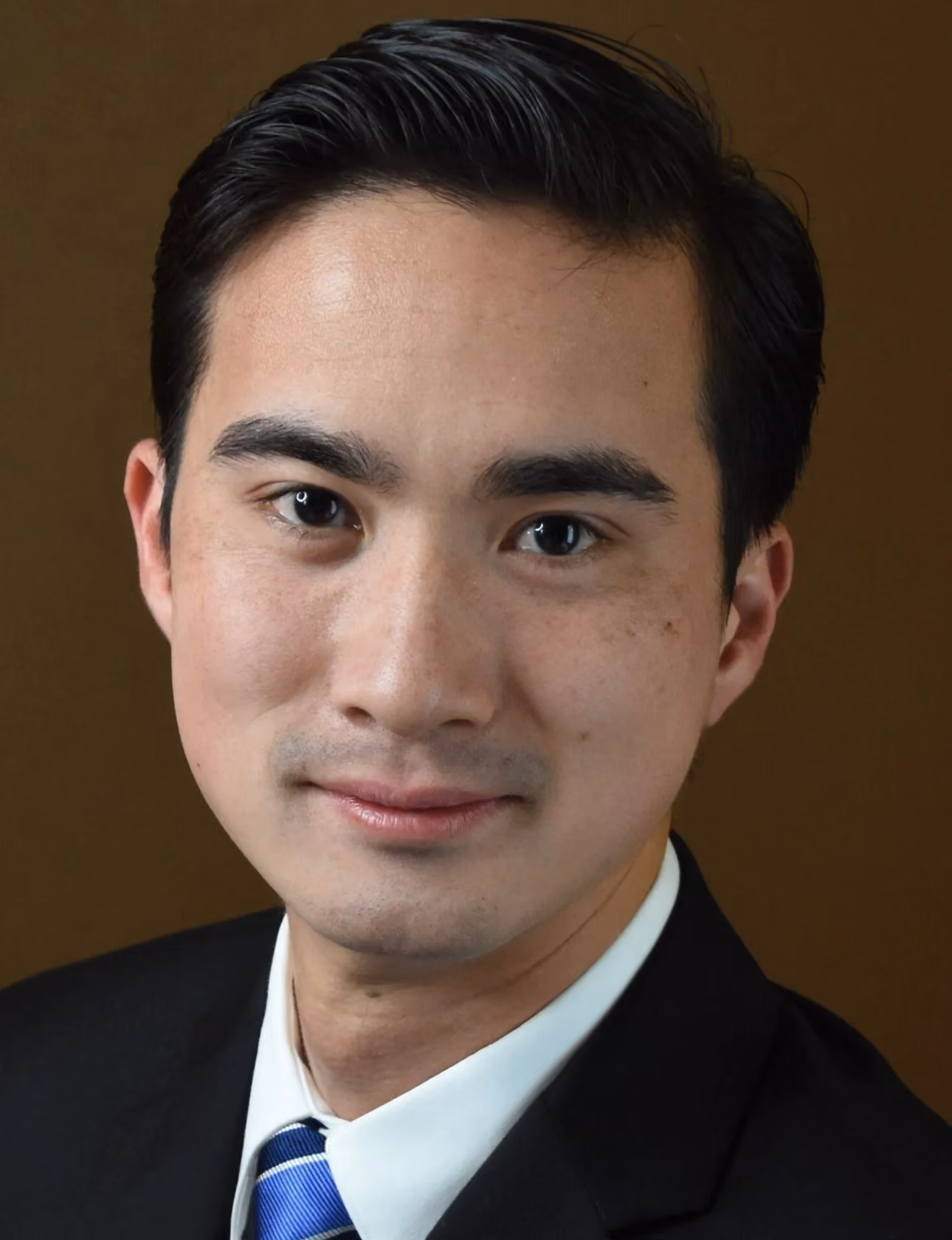 Portrait of a young man with dark hair, wearing a suit, white shirt, and blue striped tie.