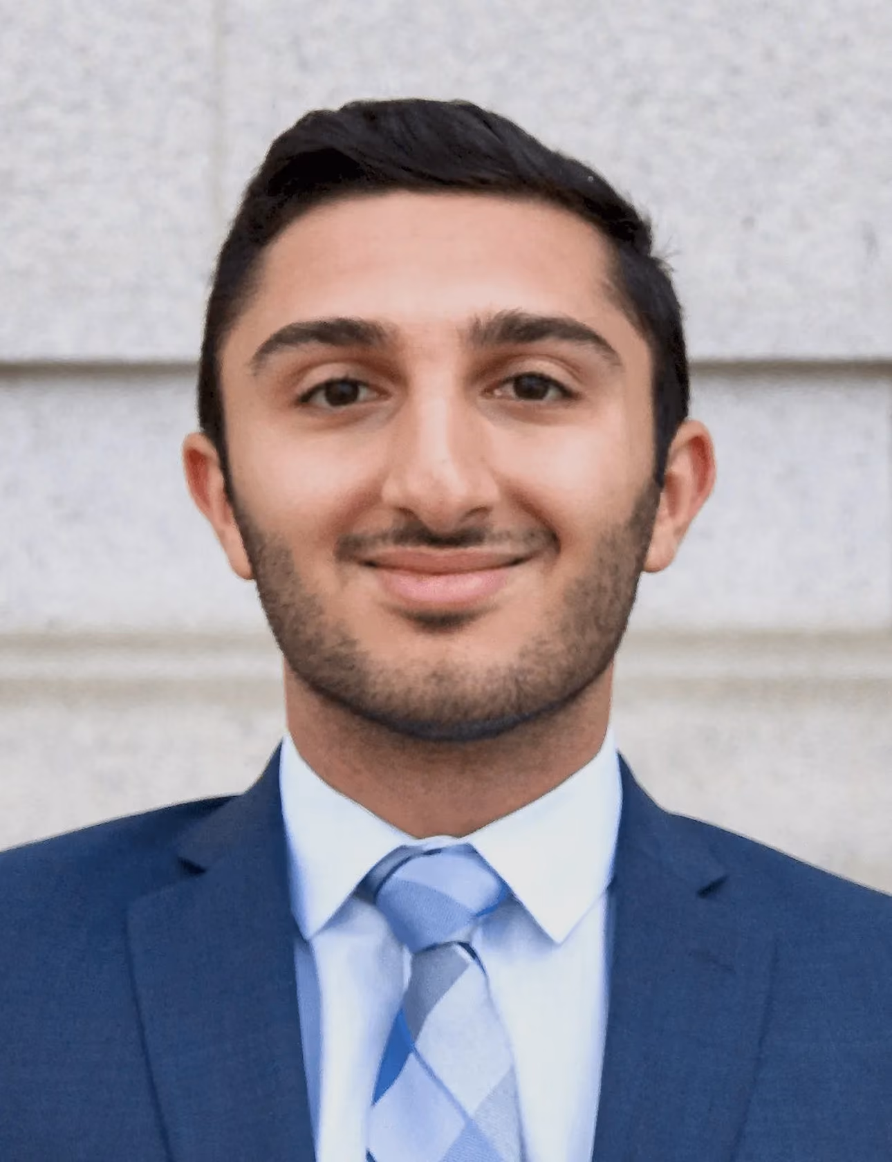 Young man with dark hair and beard wearing a blue suit and patterned tie smiling.