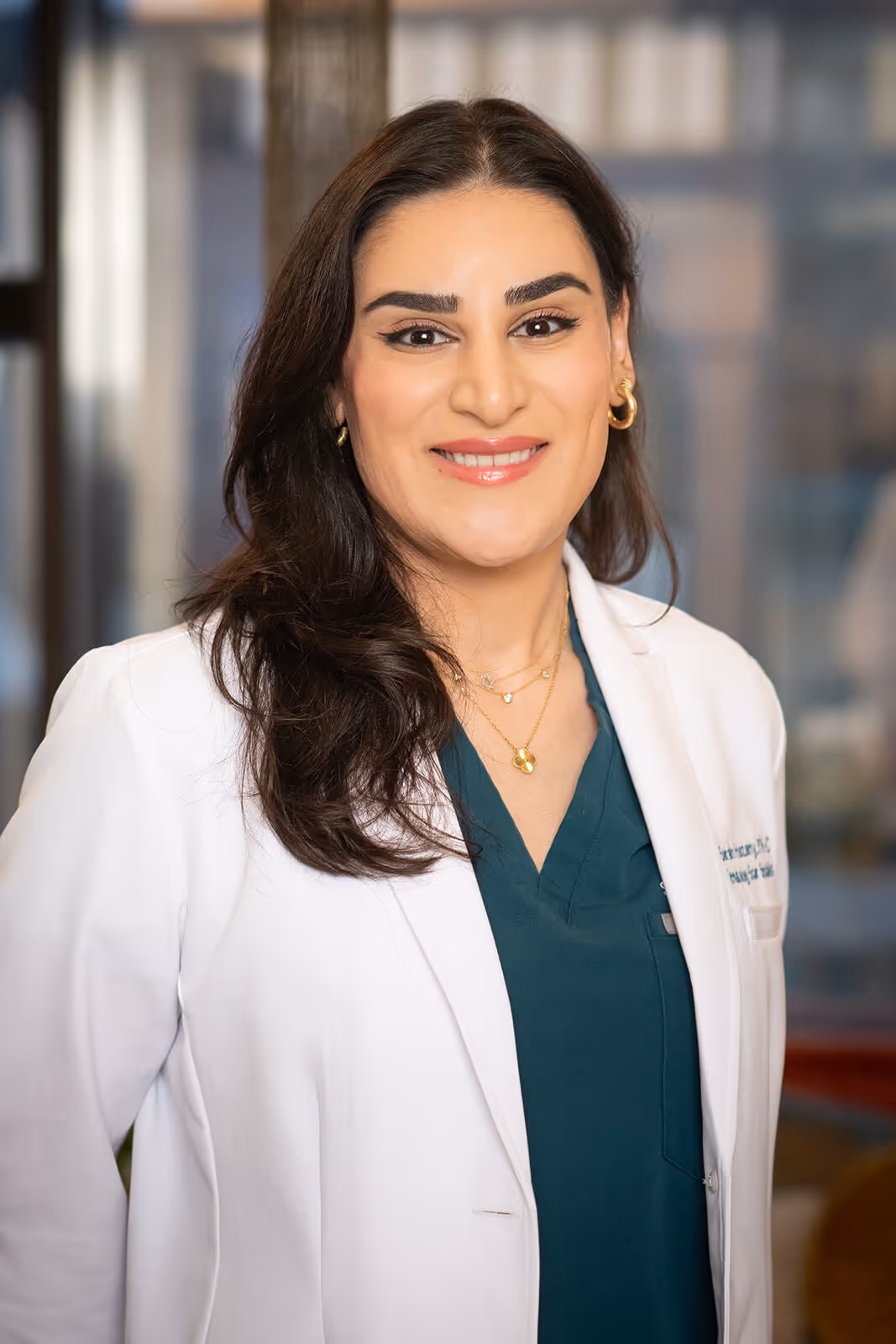Smiling woman doctor with long dark hair wearing a white coat and teal medical scrubs.