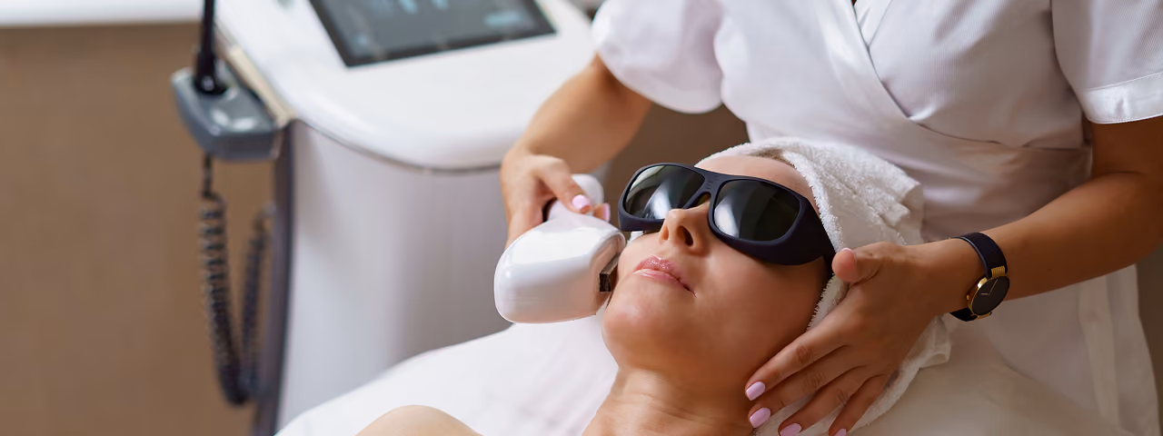 Woman wearing protective sunglasses receiving a facial laser treatment from a technician in a white uniform in a clinic.