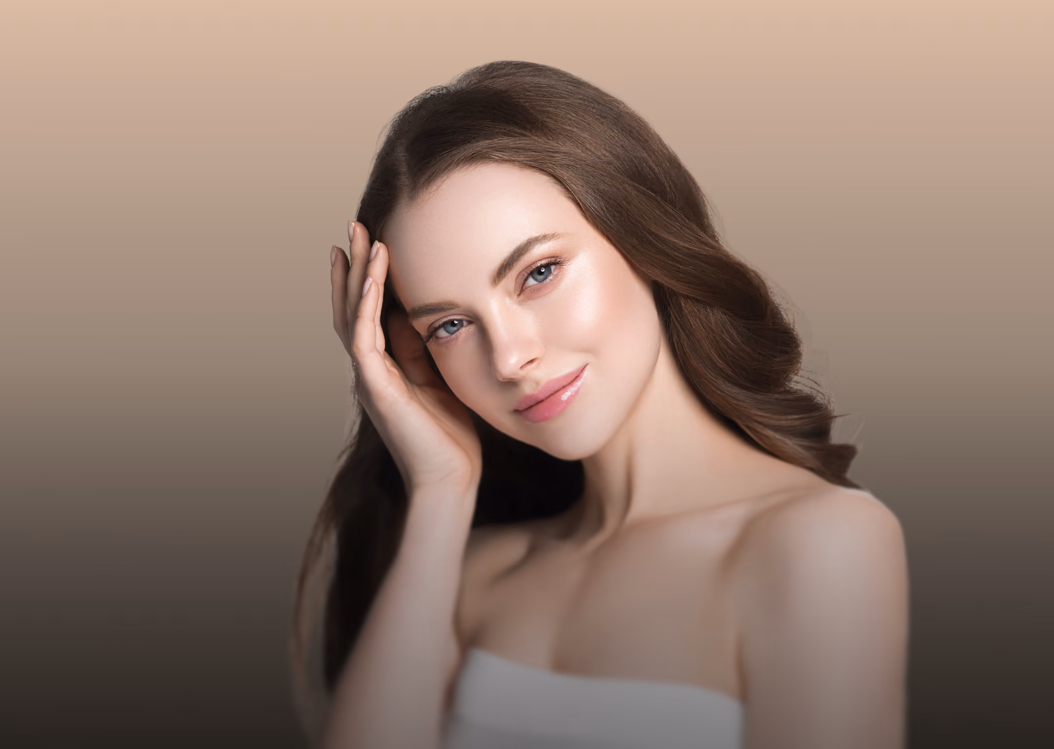 Young woman with fair skin and long brown hair touching her face and smiling softly against a beige gradient background.