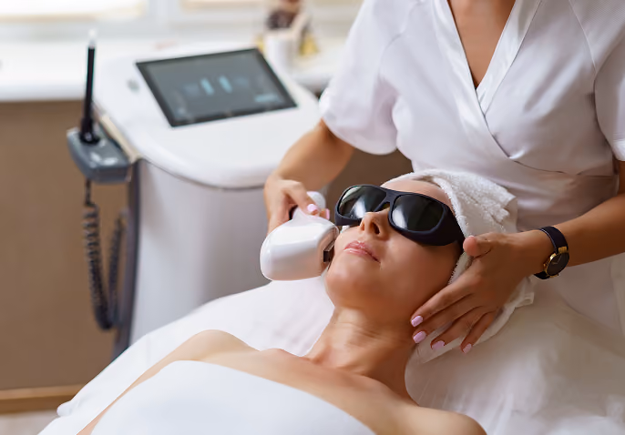 Woman wearing protective goggles receives facial laser treatment from a technician in a white uniform at a modern clinic.