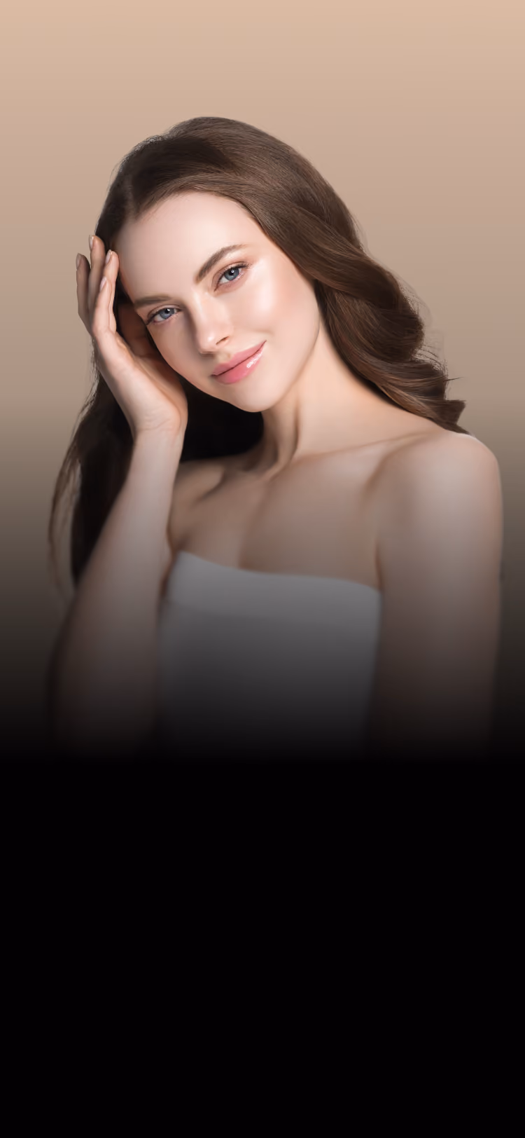 Portrait of a smiling young woman with long brown hair touching her face, wearing a strapless white top against a beige background.