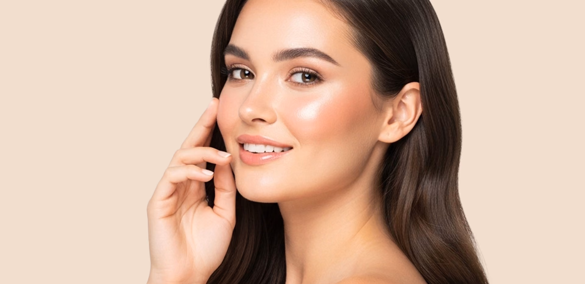Close-up of a young woman with clear skin, brown eyes, and long dark hair smiling gently while touching her face against a beige background.