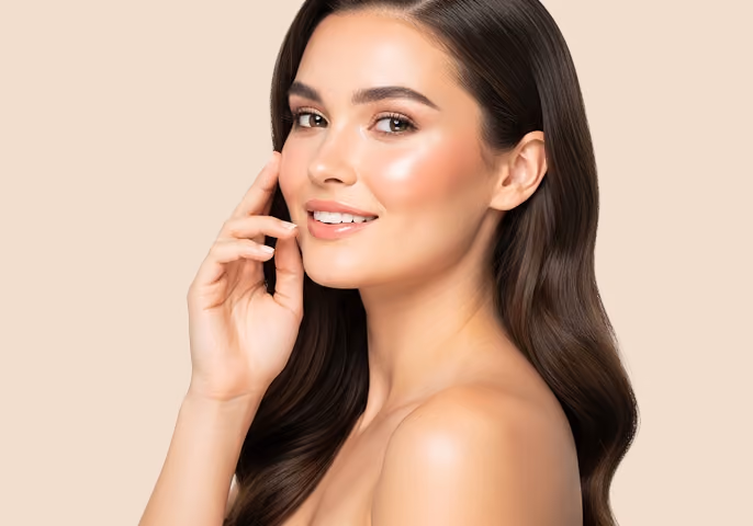 Smiling woman with long wavy brown hair touching her face, showcasing clear glowing skin against a beige background.