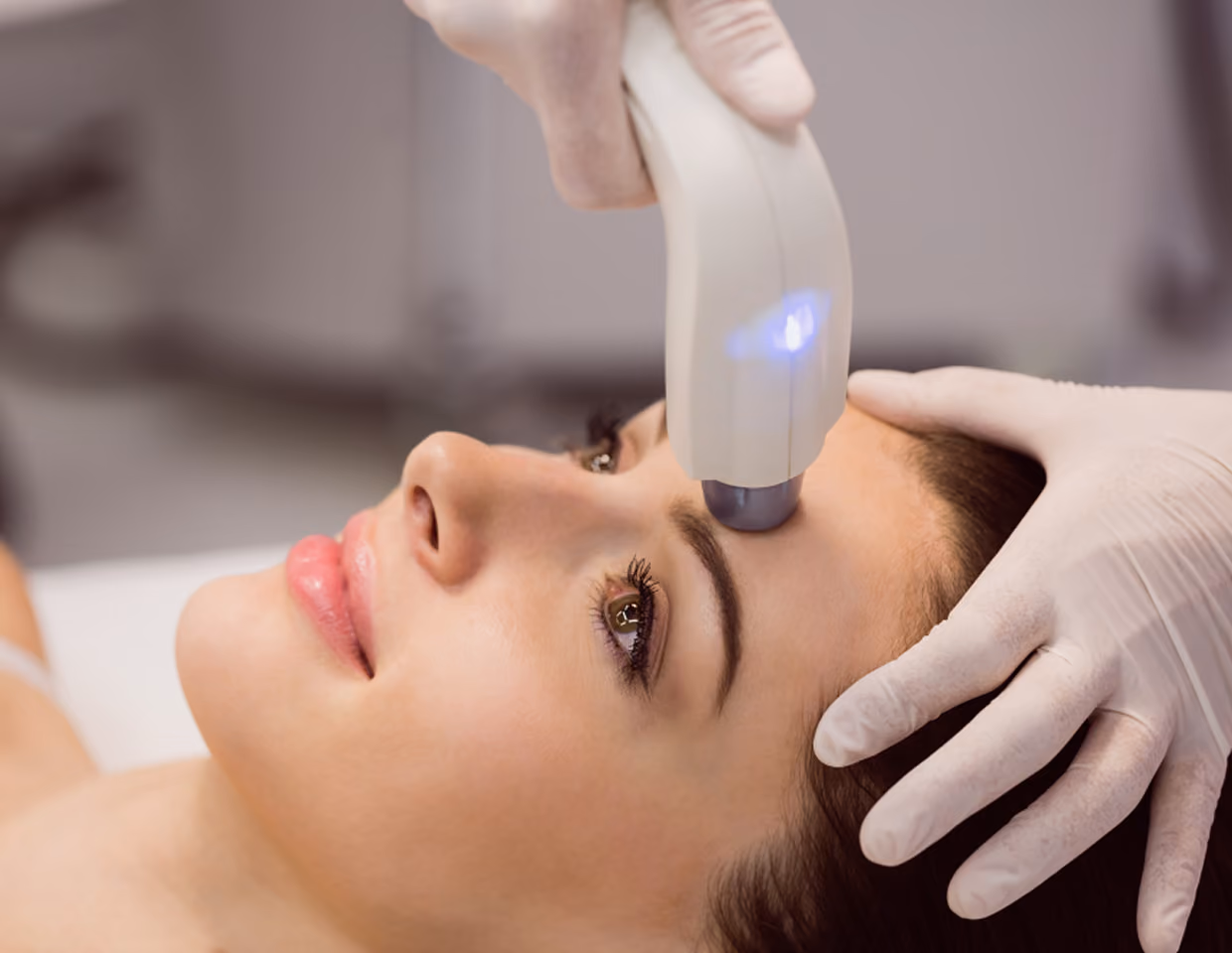 Close-up of a woman receiving a skin treatment on her forehead with a handheld device operated by a gloved professional.