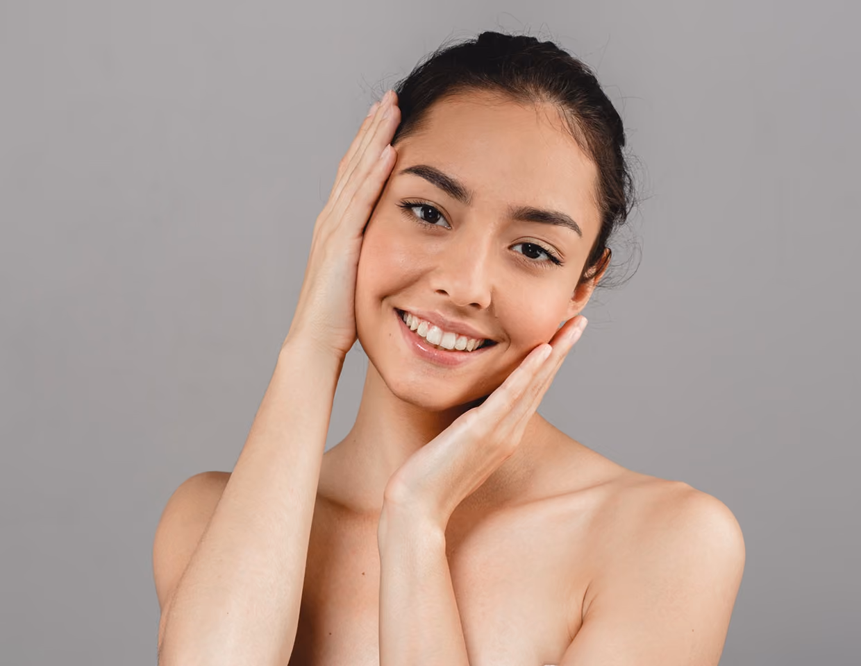 Smiling young woman with dark hair pulled back, gently touching her face with both hands against a gray background.