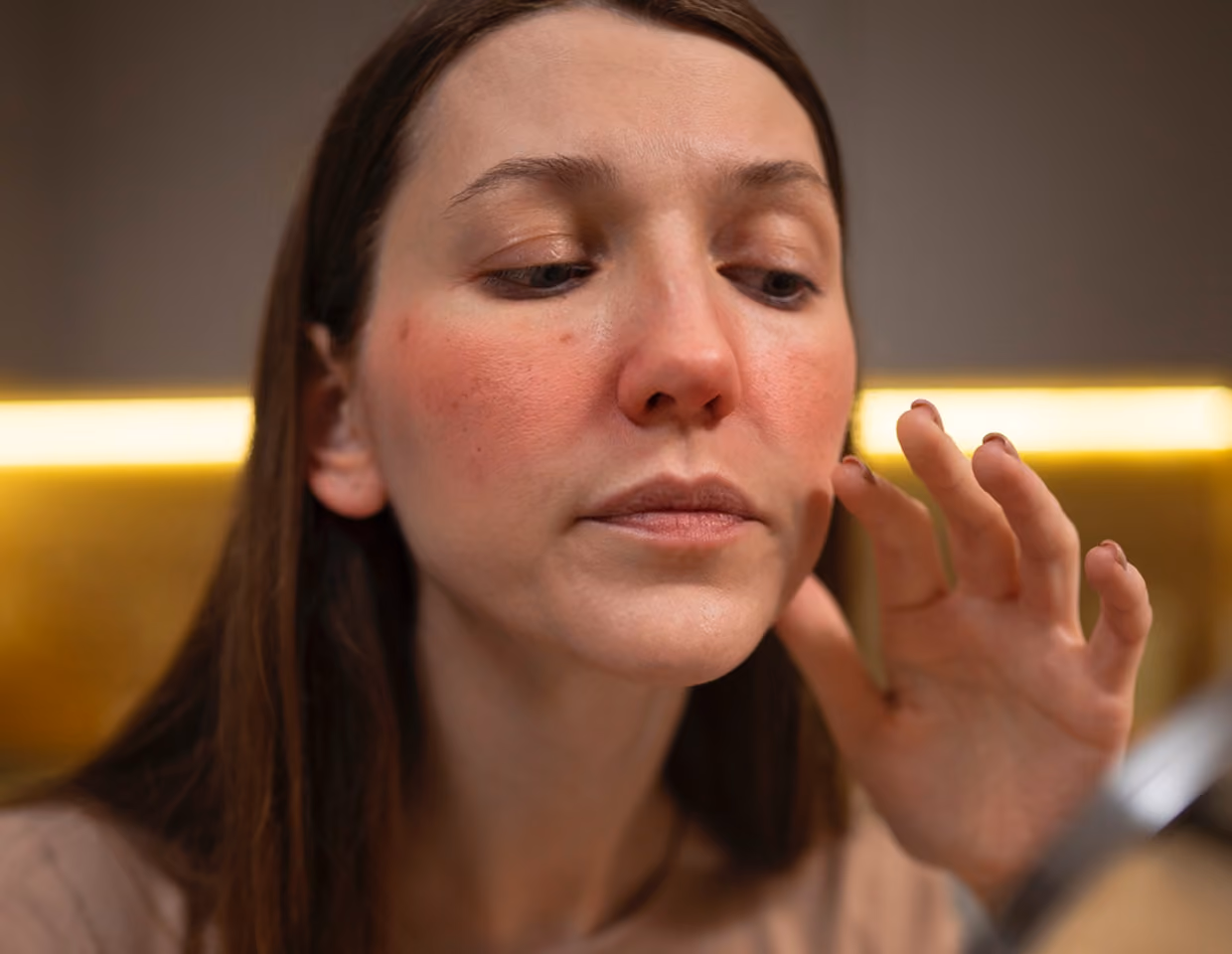 Woman with long brown hair examining her red, irritated cheeks closely in a mirror, touching her face with her hand.
