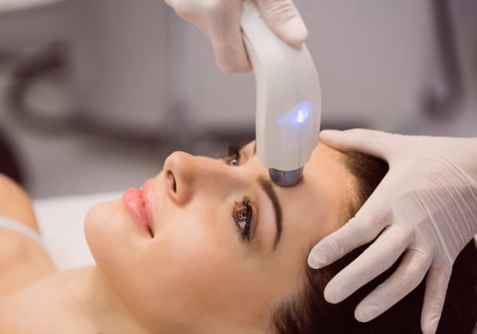 Close-up of a woman receiving a facial treatment with a handheld device on her forehead by a person wearing white gloves.